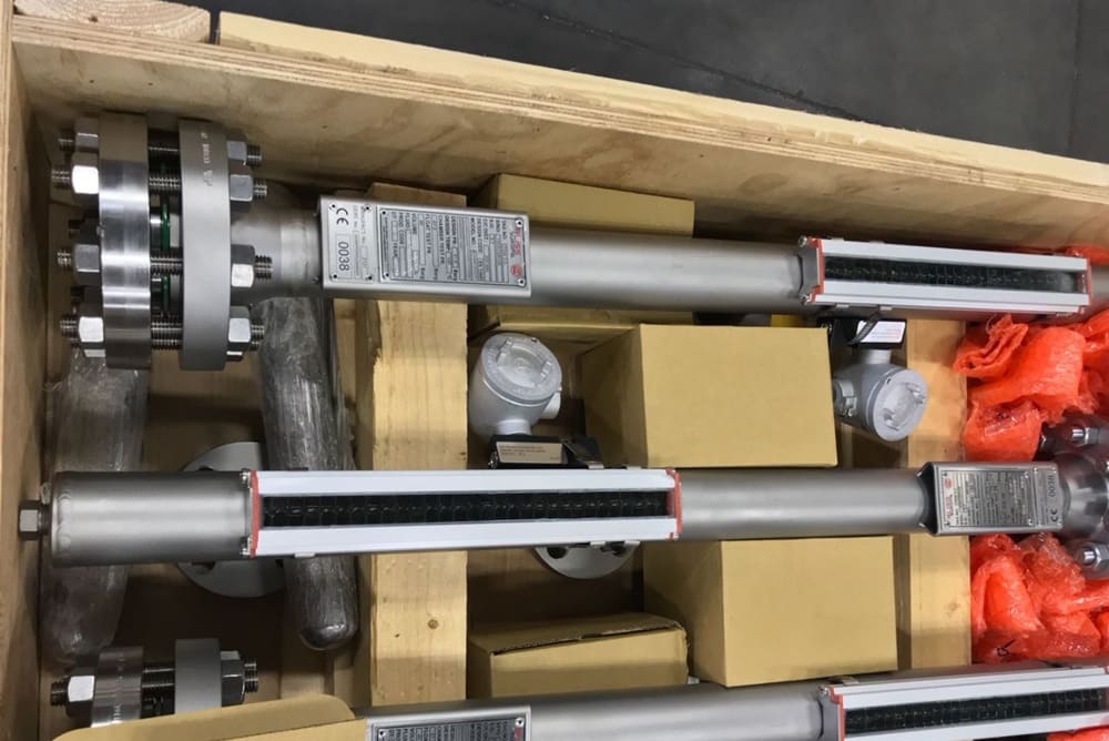 Three stainless steel industrial flow meters with flange connections packed in a wooden crate among cardboard boxes and protective foam.
