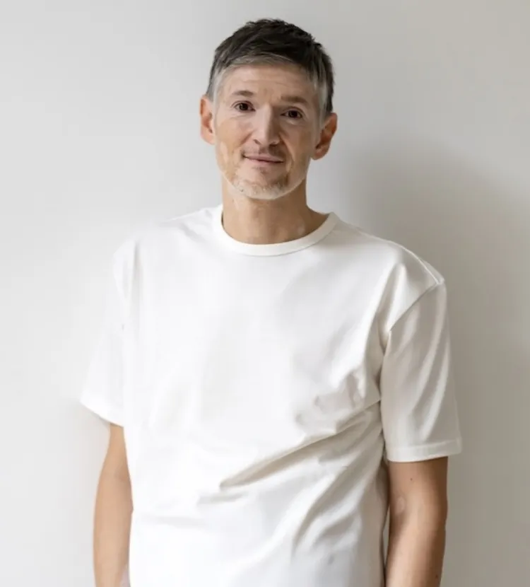 Mann mittleren Alters mit kurzen grauen Haaren trägt ein weißes T-Shirt und steht vor einer weißen Wand.