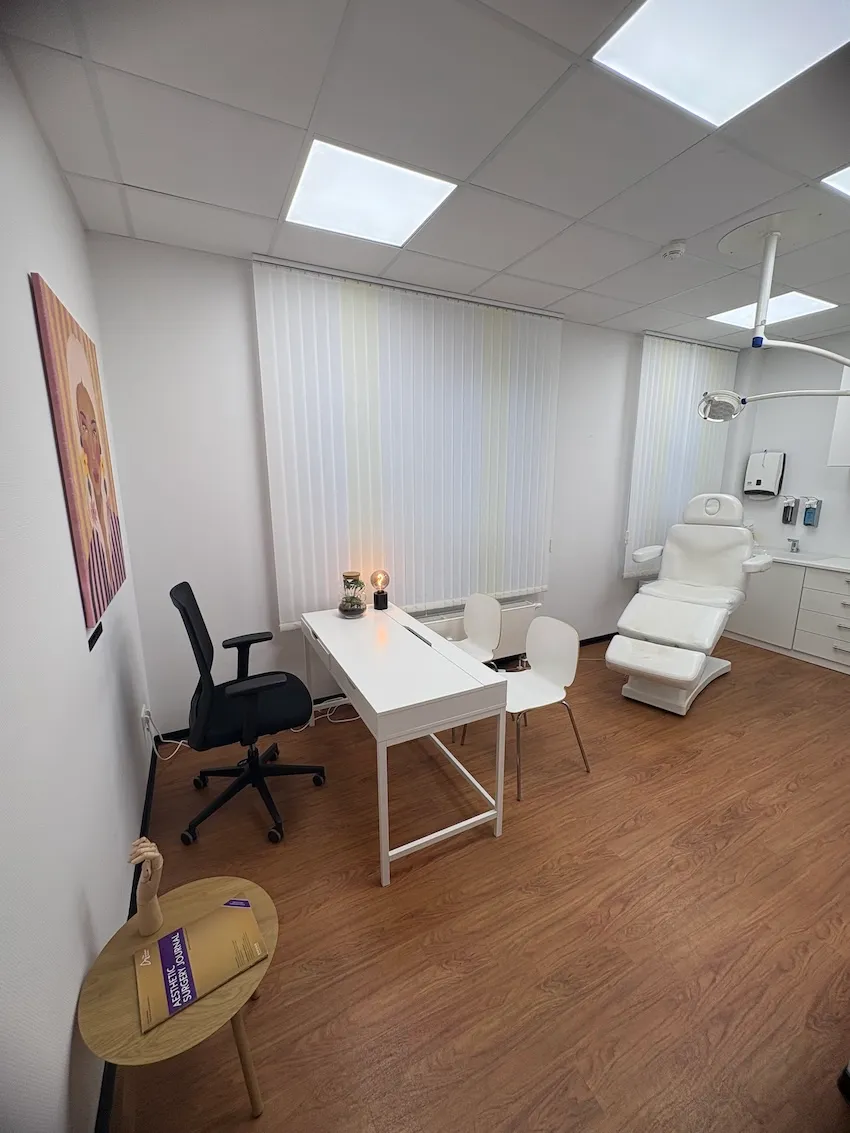 Behandlungszimmer mit einem weißen Behandlungssessel, einem Schreibtisch mit zwei weißen Stühlen und einem schwarzen Bürostuhl, hellen Vorhängen und Holzboden.