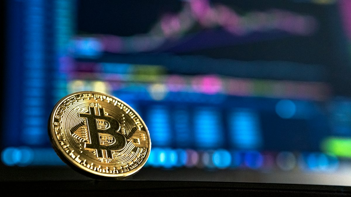 A gold bitcoin coin in front of a blurred trading screen.