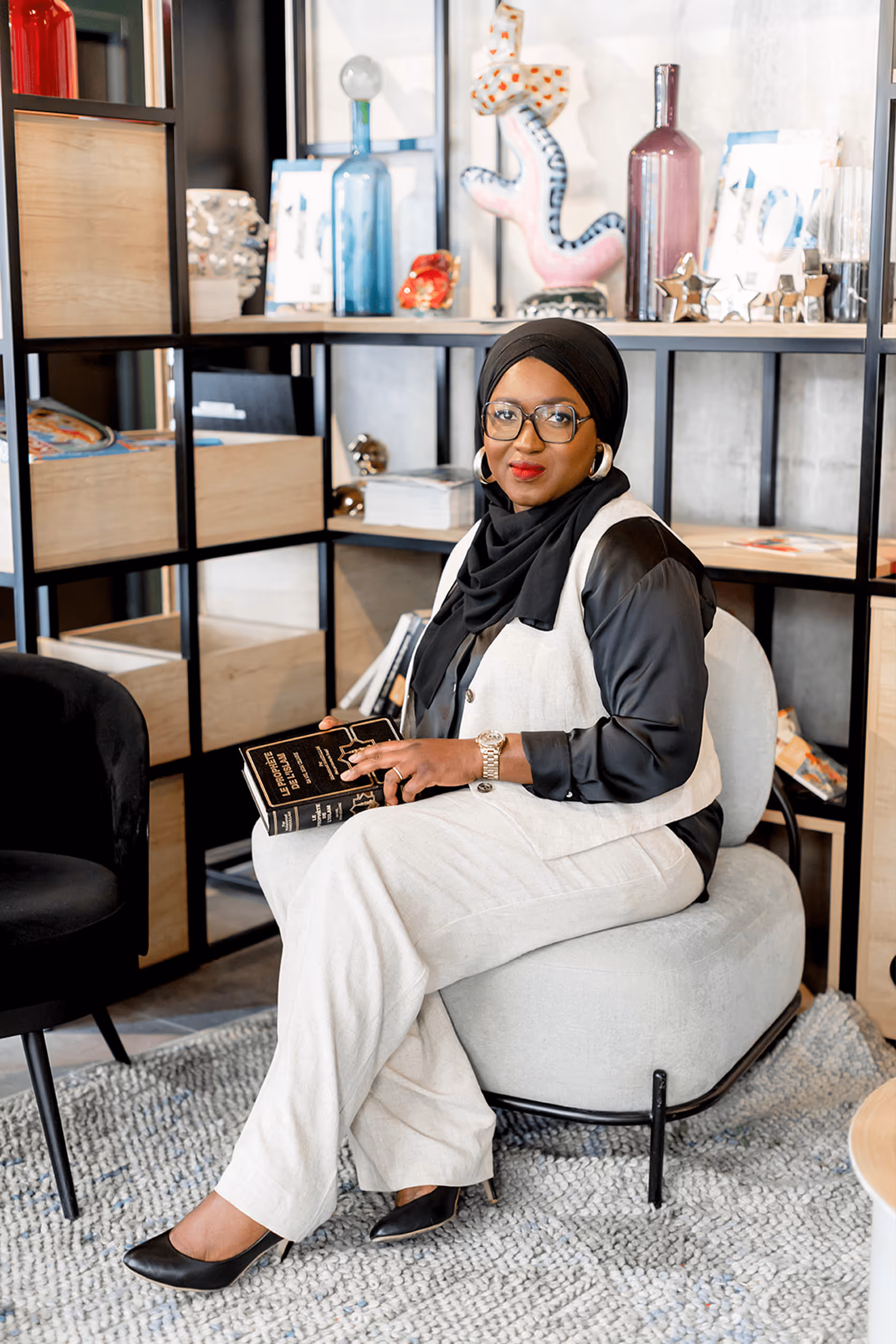 Tity Diomandé, une femme élégante avec des lunettes et un ensemble noir et beige, est assise dans un fauteuil moderne avec une bibliothèque en arrière-plan. Elle tient un livre dans les mains et regarde l’objectif avec un léger sourire.