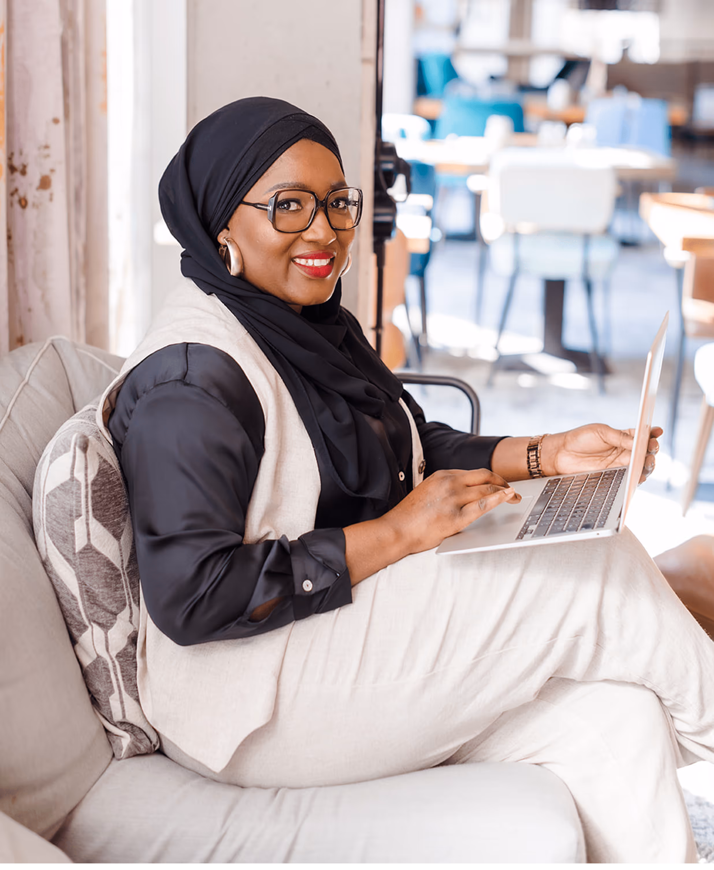 Tity, coach musulmane, assise sur un canapé avec un ordinateur portable sur les genoux, souriante et détendue dans un espace de coworking lumineux.