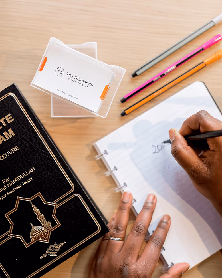 Main d'une femme écrivant '202...' dans un carnet, avec un exemplaire du livre "Le Prophète de l'islam", des stylos colorés et des cartes de visite posées sur une table en bois clair.