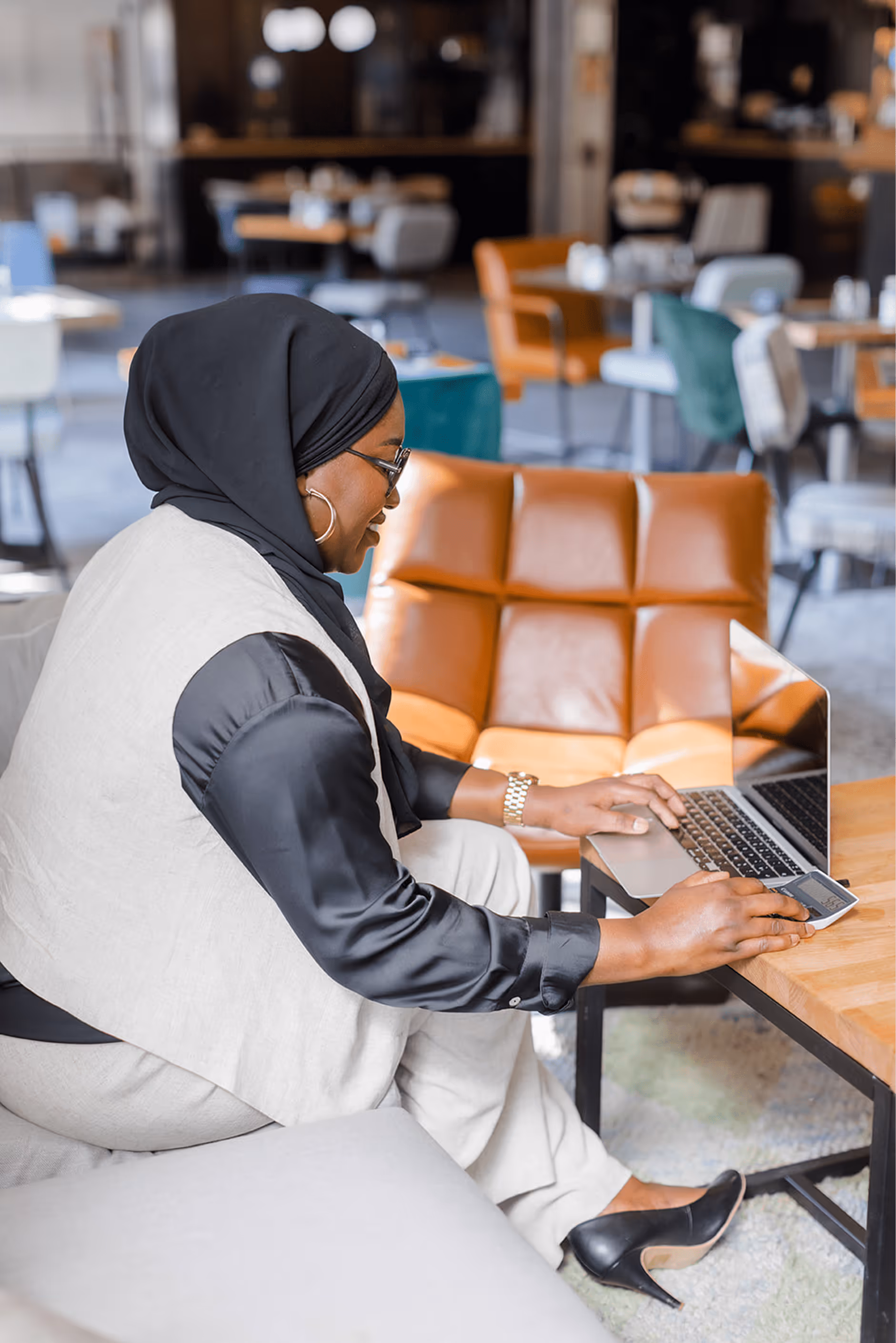 Tity, coach musulmane, concentrée sur son ordinateur portable dans un espace de travail moderne, assise sur une banquette face à une table basse.