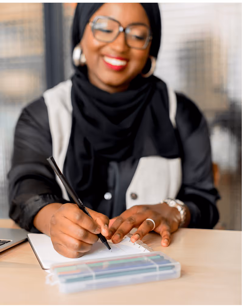 Tity, coach musulmane, souriante et concentrée, en train d’écrire dans un carnet sur un bureau, avec une trousse de stylos colorés à portée de main.
