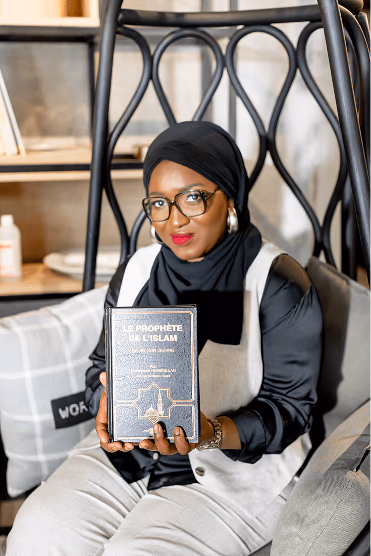 Tity, coach musulmane, assise et souriante, tenant un exemplaire du livre 'Le Prophète de l’Islam' dans un fauteuil au design moderne.