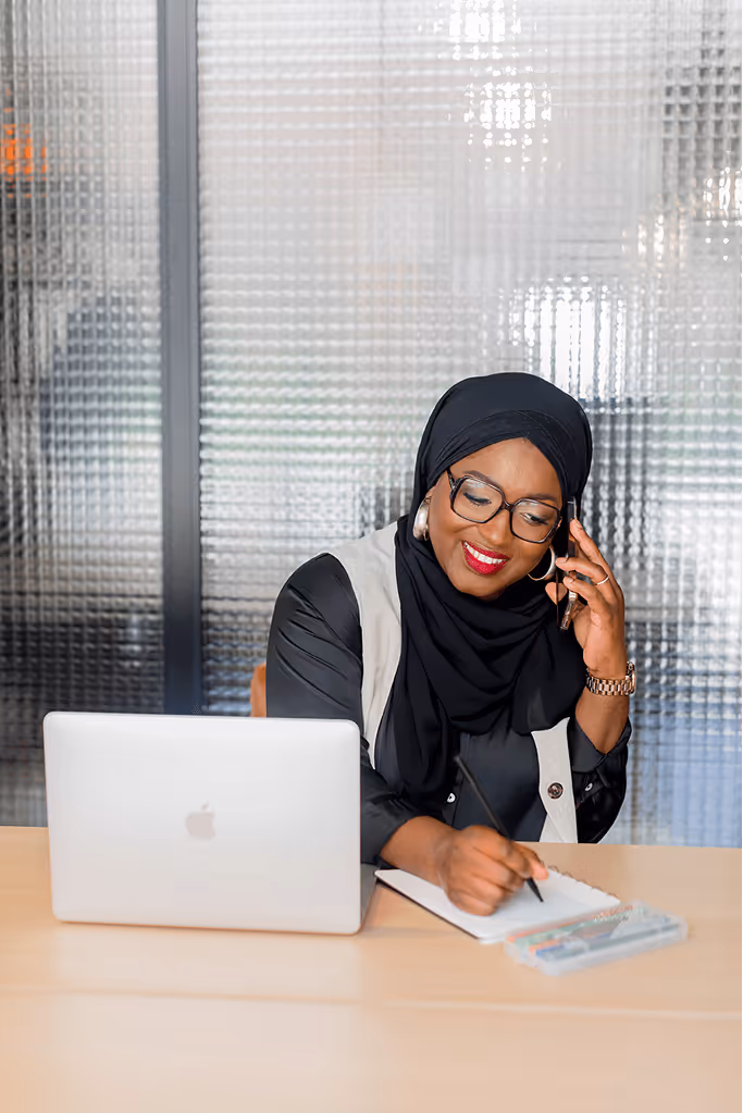 Tity, coach musulmane, souriante au téléphone tout en prenant des notes devant son ordinateur portable, dans un cadre professionnel lumineux.