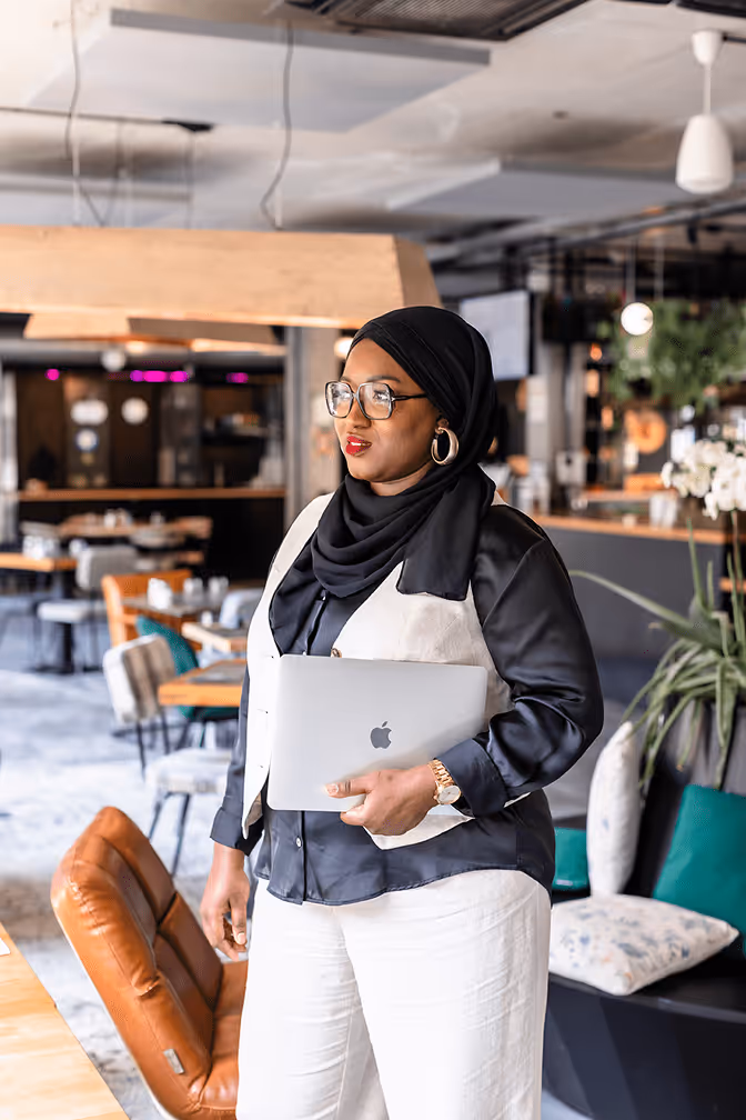Tity, coach musulmane, debout avec un ordinateur portable à la main, regard déterminé dans un espace de coworking moderne et élégant.