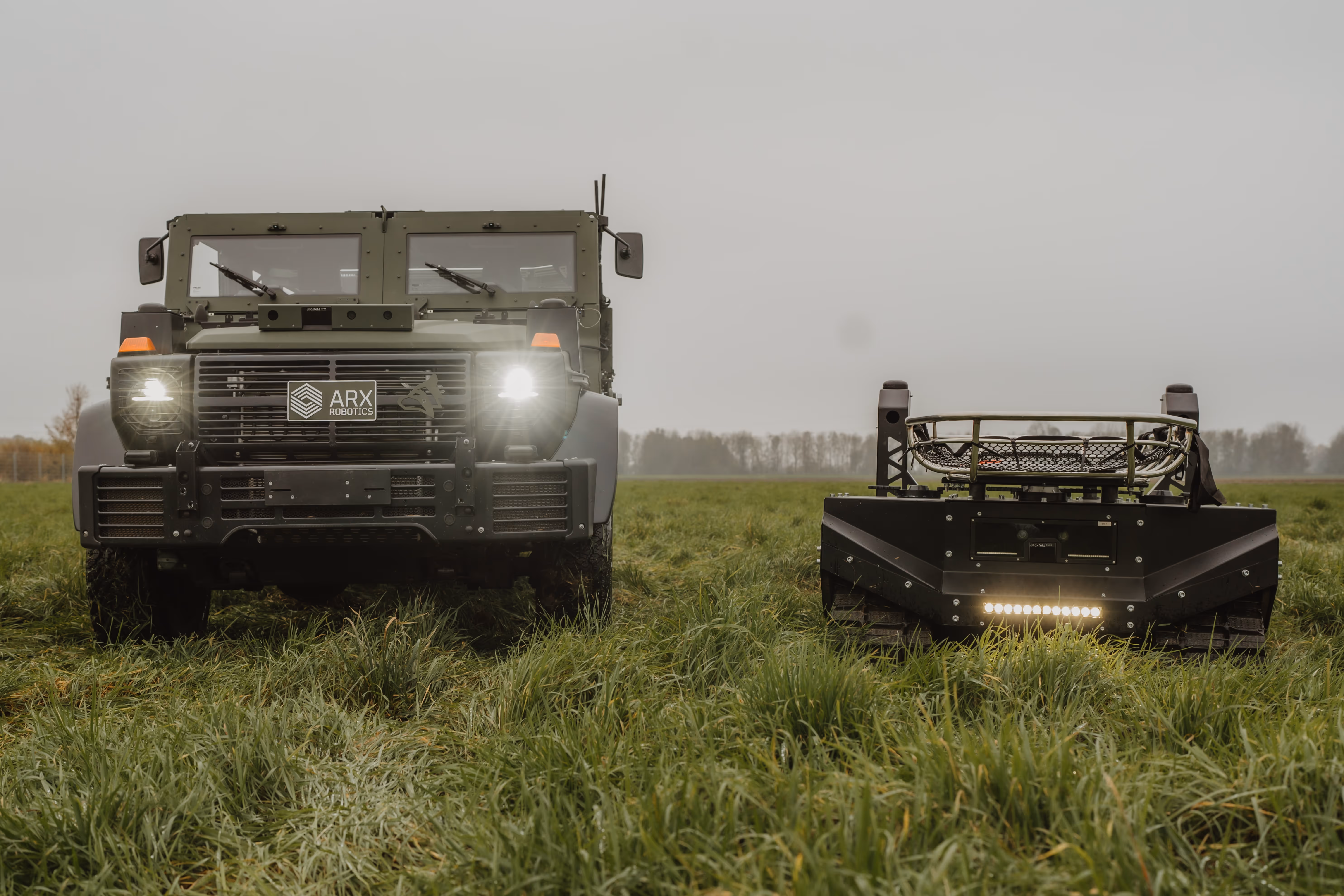 Front view of two autonomous robotic vehicles on grass, one resembling a military truck with headlights on and ARX Robotics logo, the other a smaller tracked robot with lights.