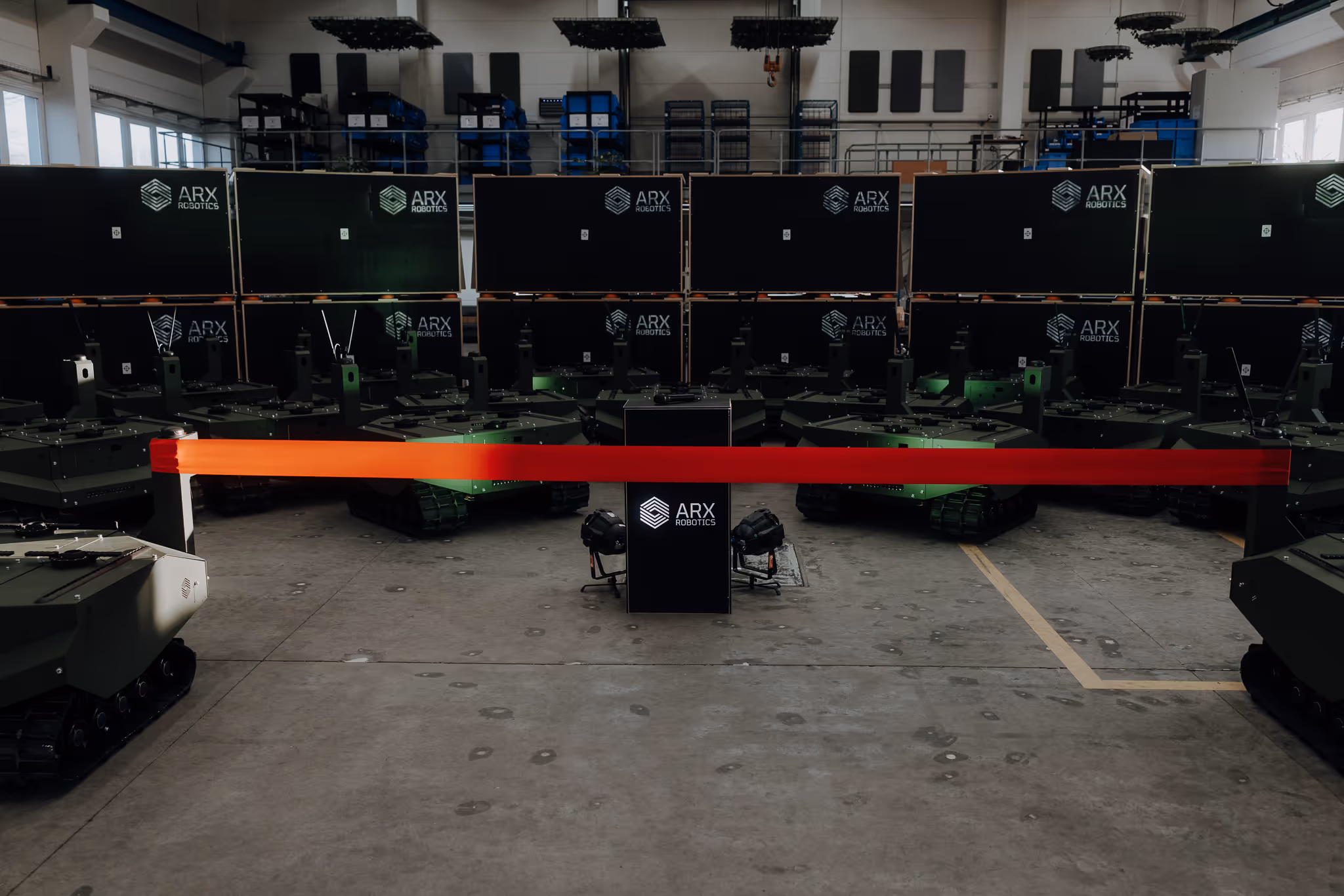 Indoor warehouse showing a lineup of military-style robotic vehicles with ARX Robotics logos and a red barrier tape in front.