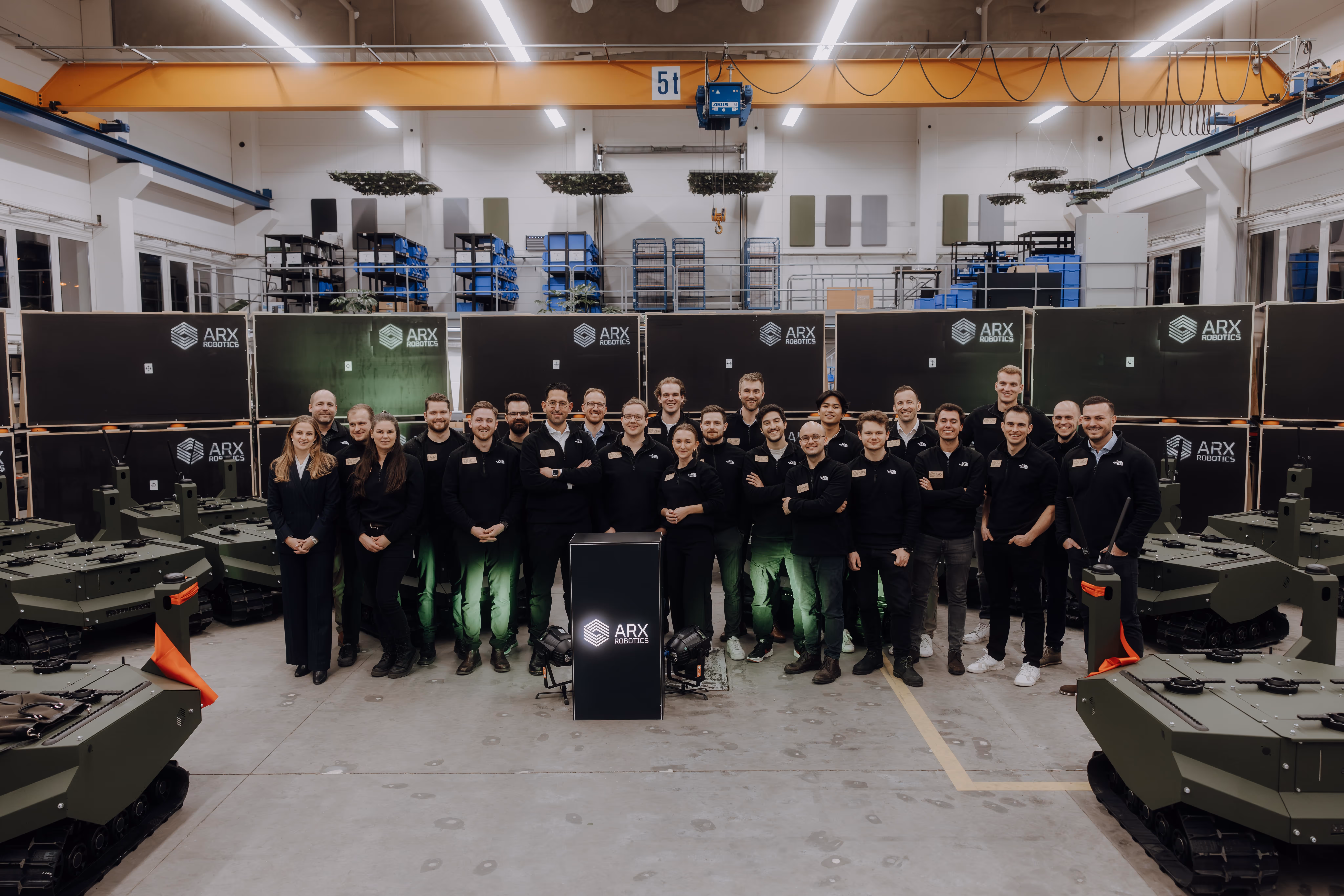 Group photo of ARX Robotics team standing inside a warehouse surrounded by green robotic vehicles and black crates with ARX Robotics logos.