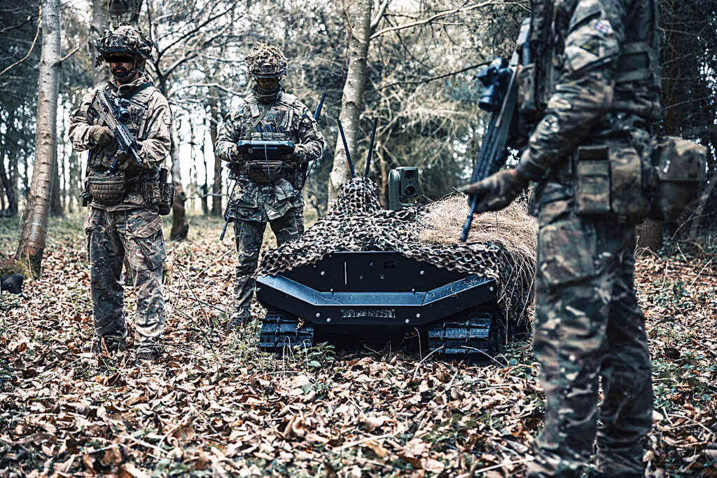 Three soldiers in camouflage gear operating a small tracked unmanned vehicle covered with camouflage netting in a forest with fallen leaves.
