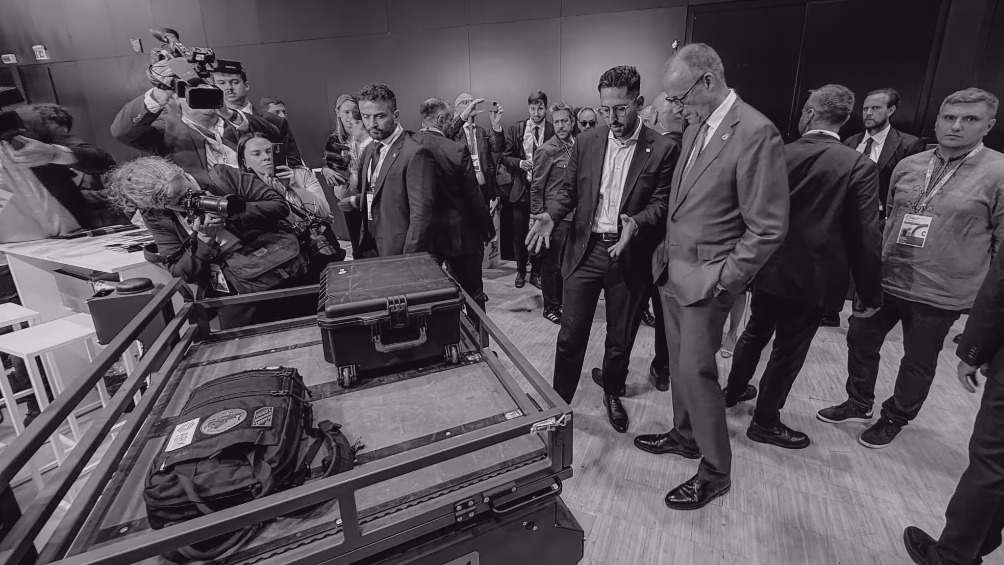 Group of people, including photographers and two men in suits, gathered around a cart carrying two closed cases.
