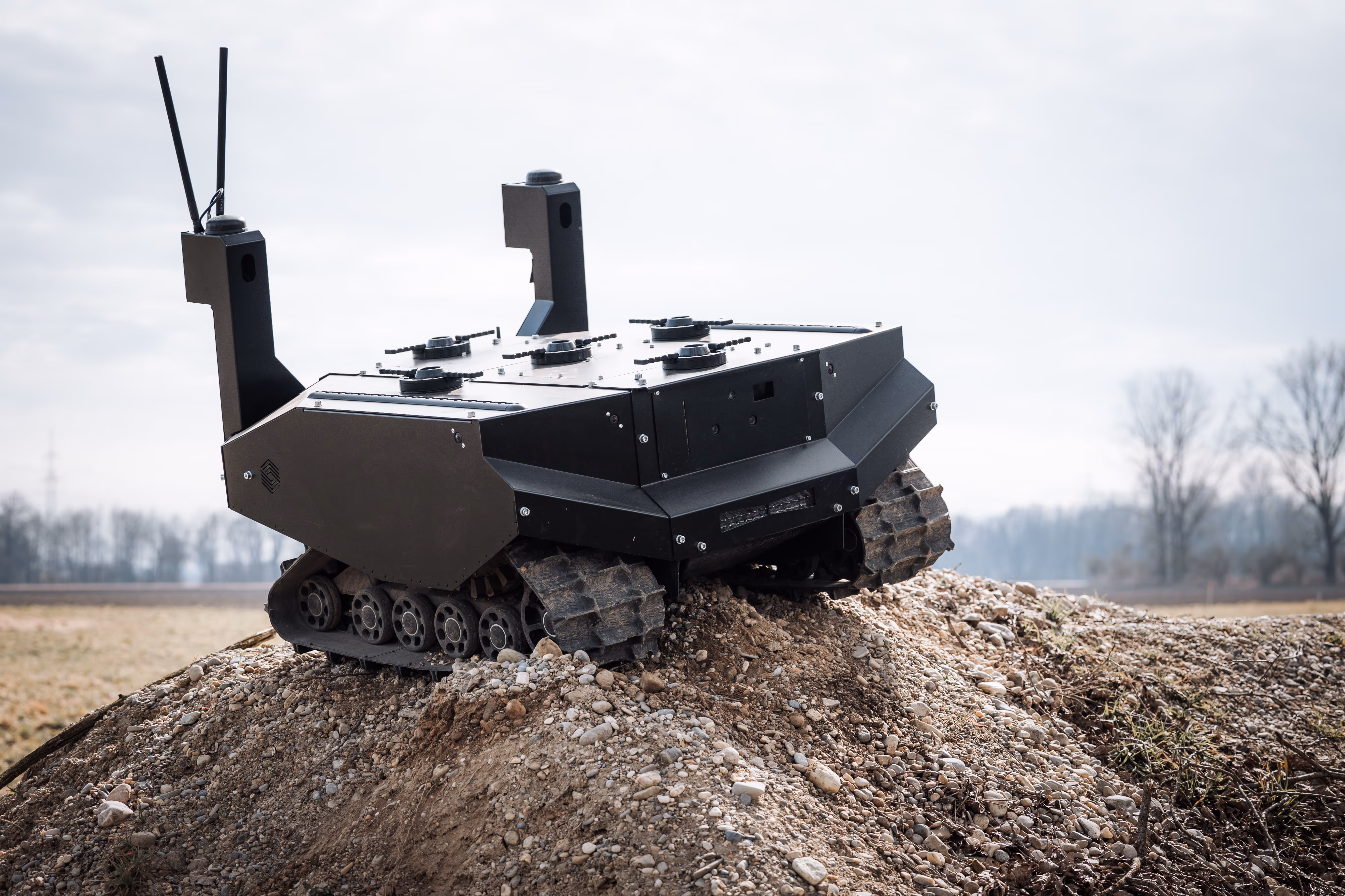 Black robotic vehicle with tank-like tracks positioned on a rocky dirt mound in an outdoor setting.