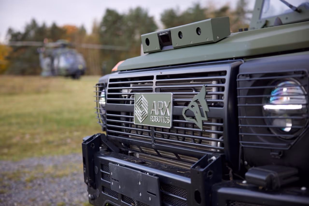 Front grill of an ARX Robotics military vehicle with mounted sensors, a headlight, and a blurred helicopter in the background.