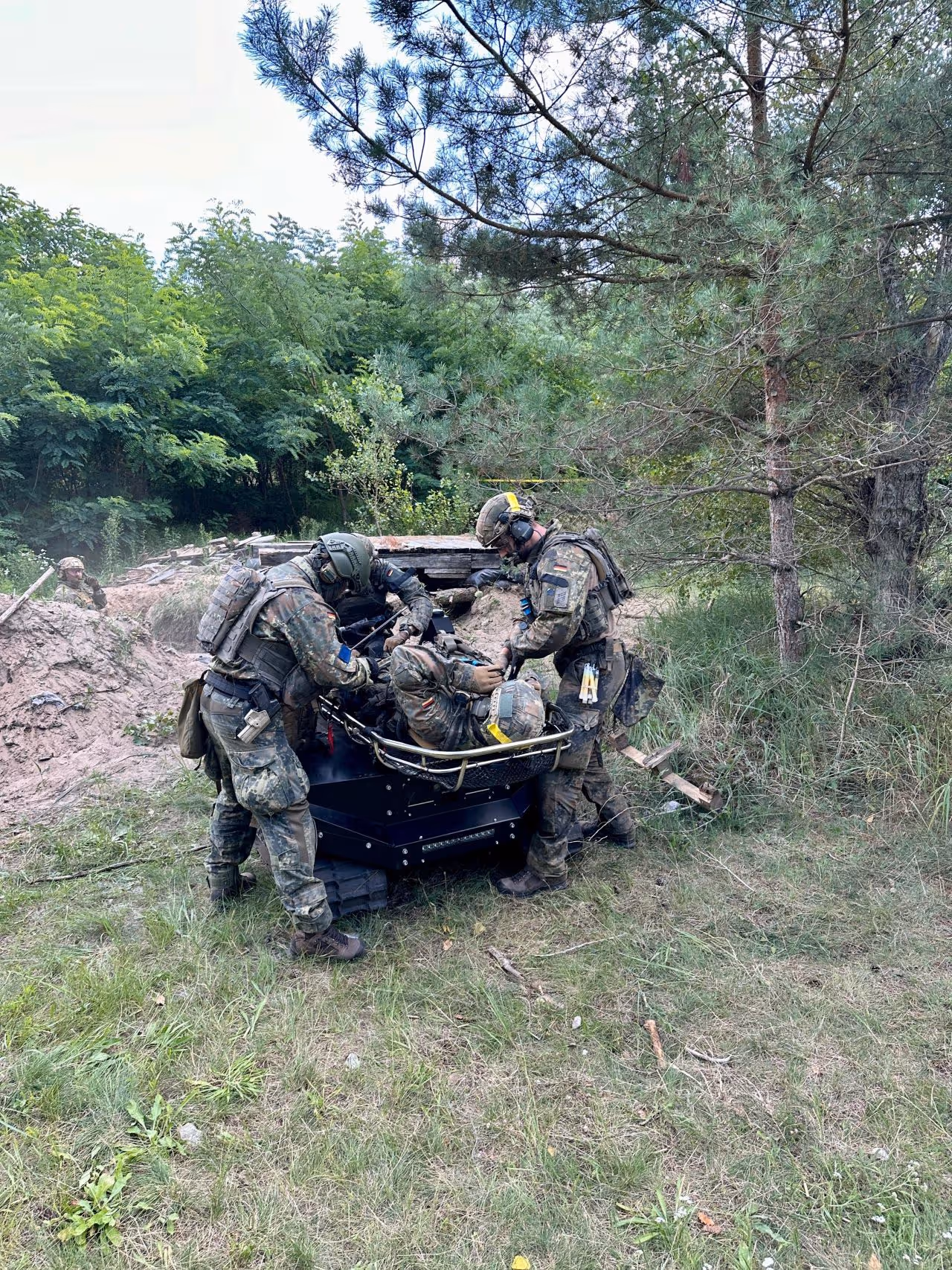 Two soldiers in camouflage gear assist a wounded comrade lying on a stretcher mounted on a robotic tracked vehicle in a forested area.