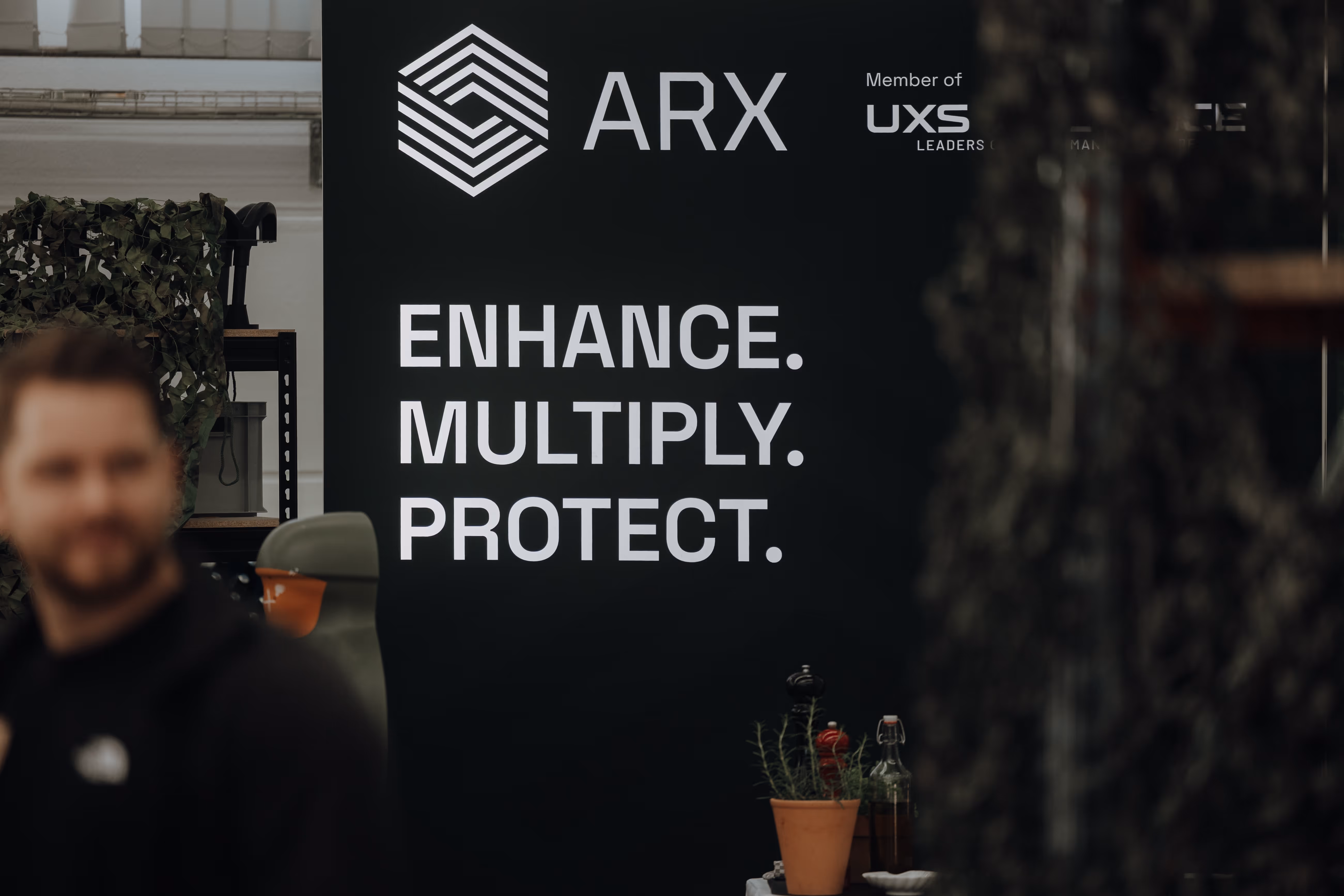 Black sign with ARX logo and words Enhance. Multiply. Protect. with blurred man in foreground.
