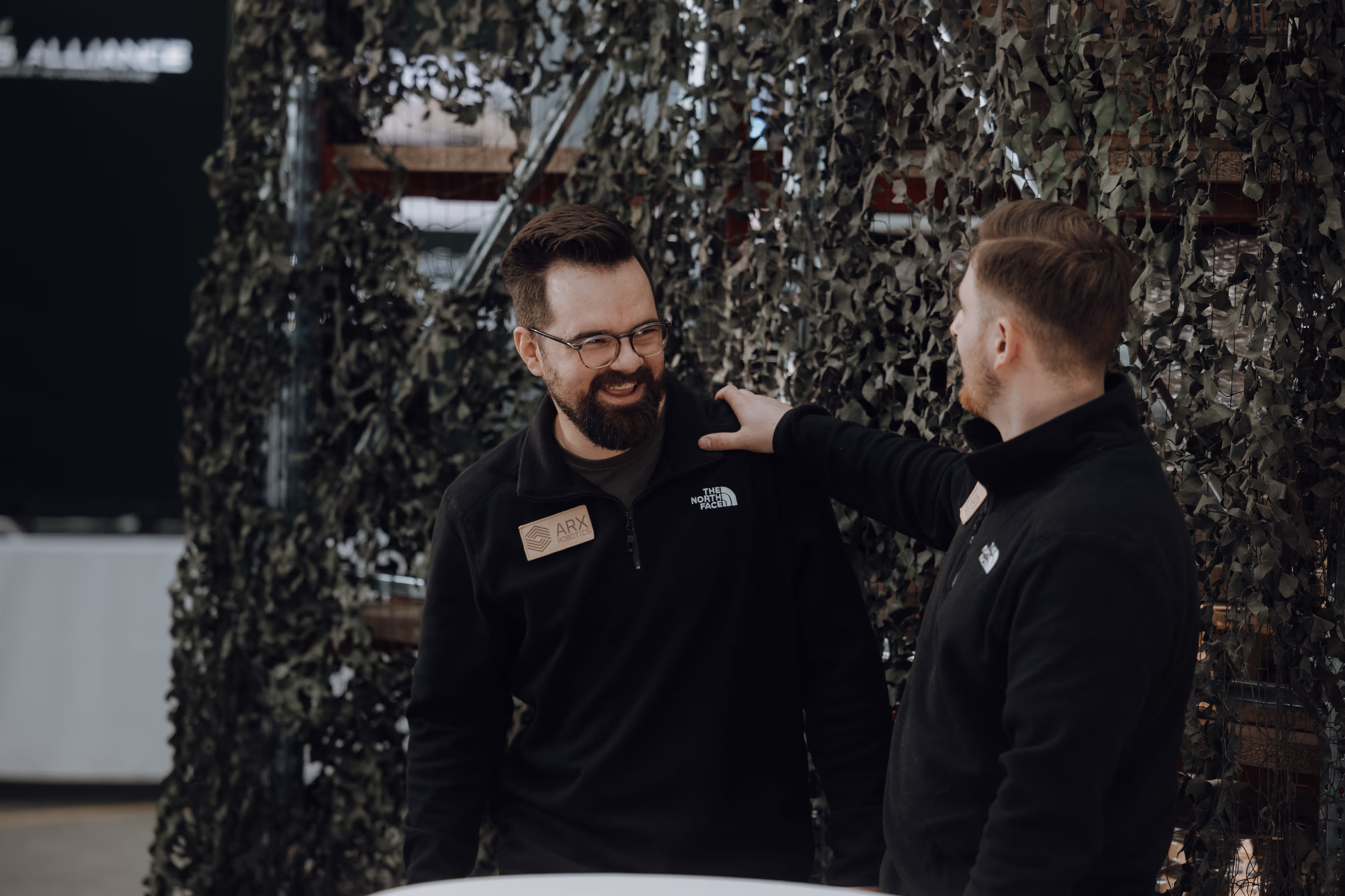 Two men wearing black North Face jackets smiling and conversing, one with glasses and beard, the other with hand on his shoulder, standing in front of a green leafy backdrop.