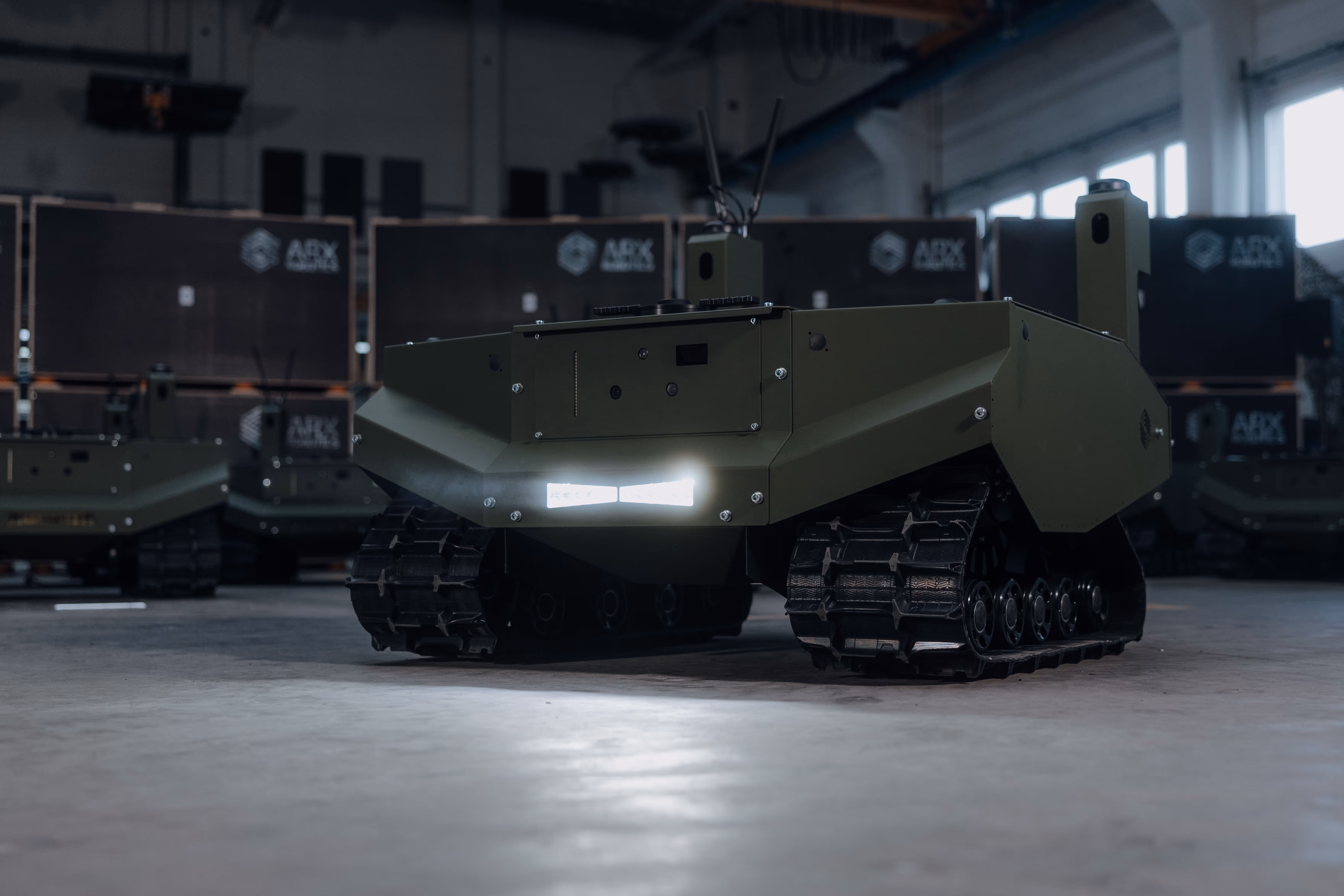 Low-profile military robot with tank tracks and antenna standing indoors on a concrete floor.