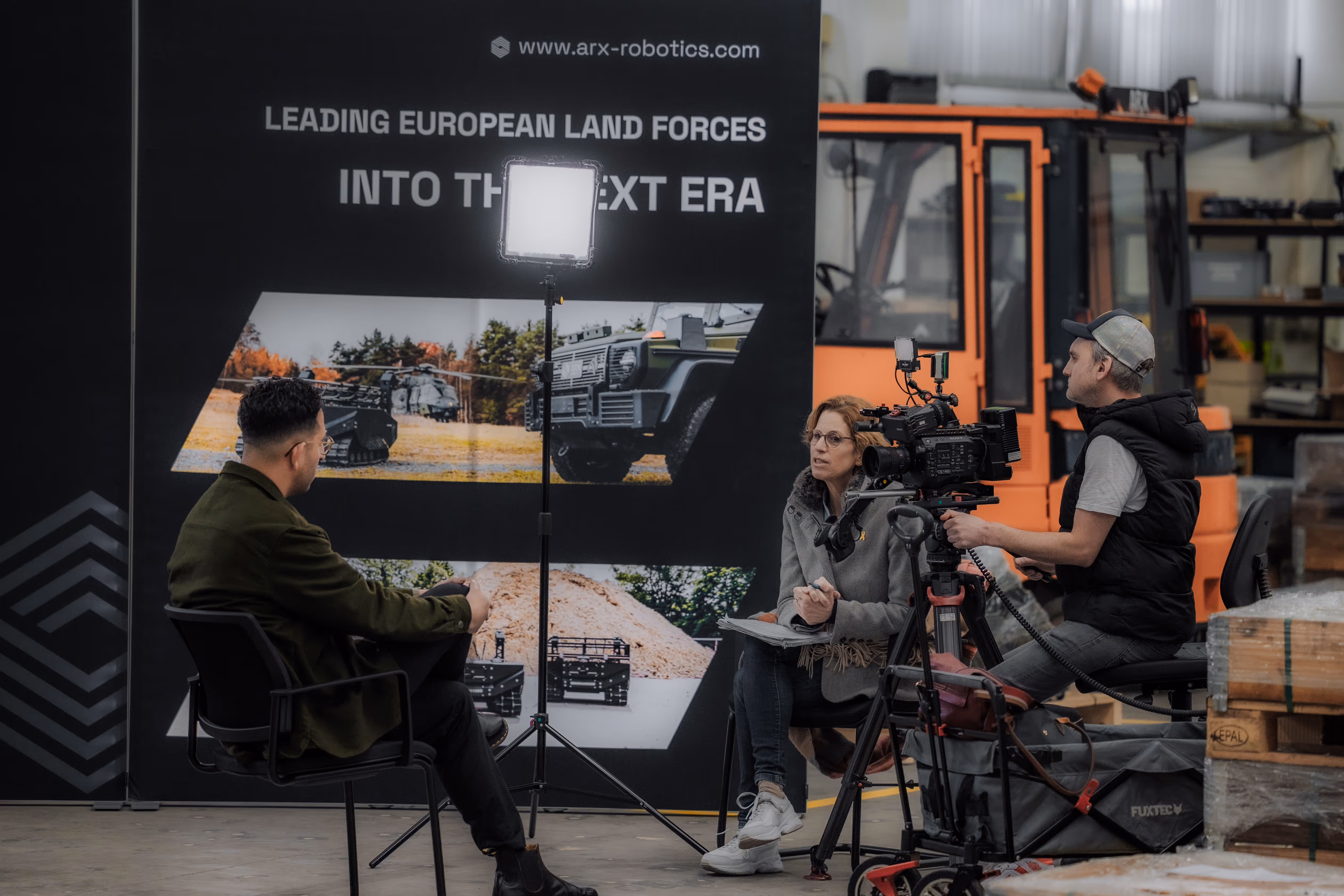 Three people in an indoor industrial setting: one man seated for an interview, a woman holding a notepad, and a man operating a video camera with lighting equipment.