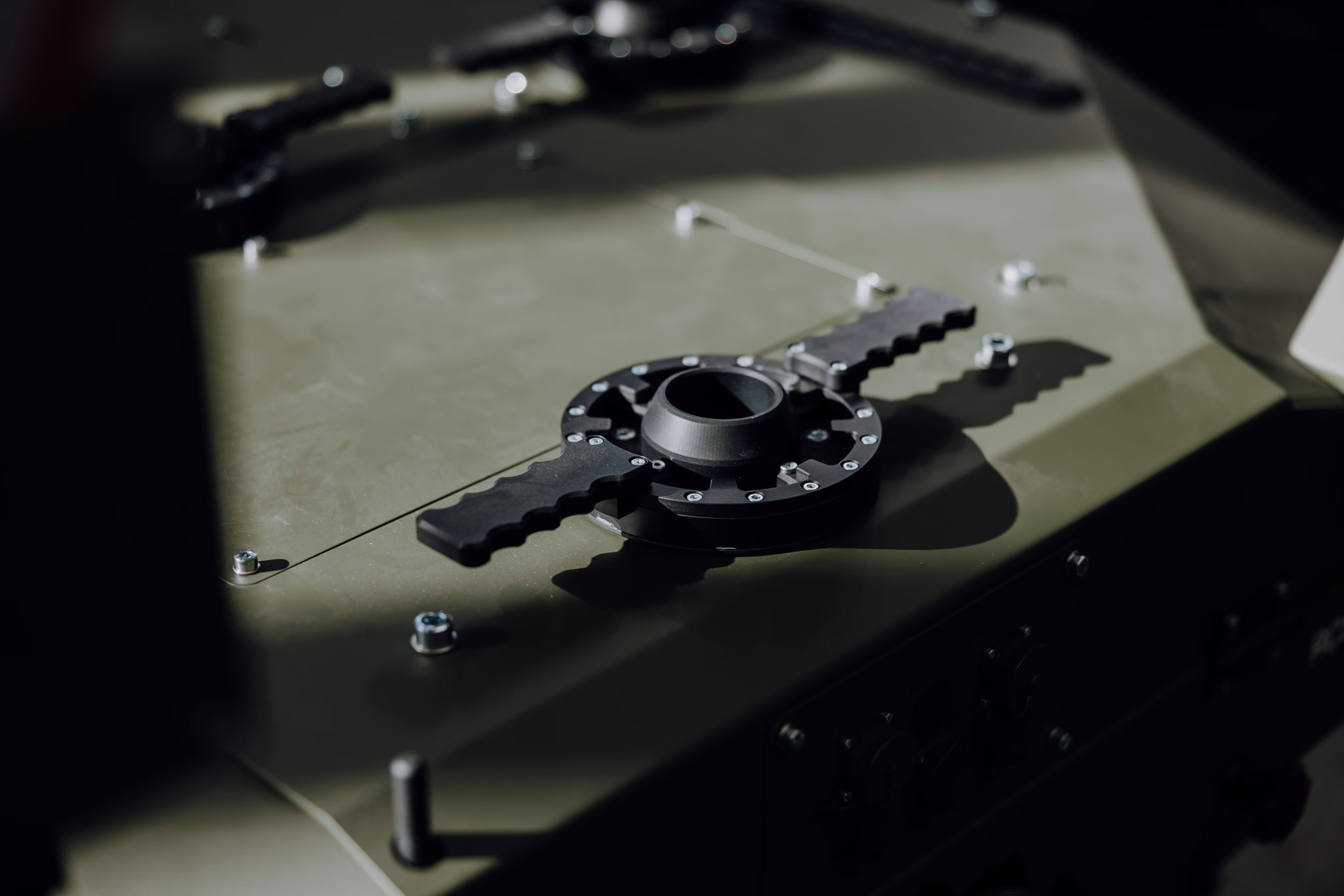 Close-up of a black circular mechanical attachment with two textured handles on a green metallic surface with screws.
