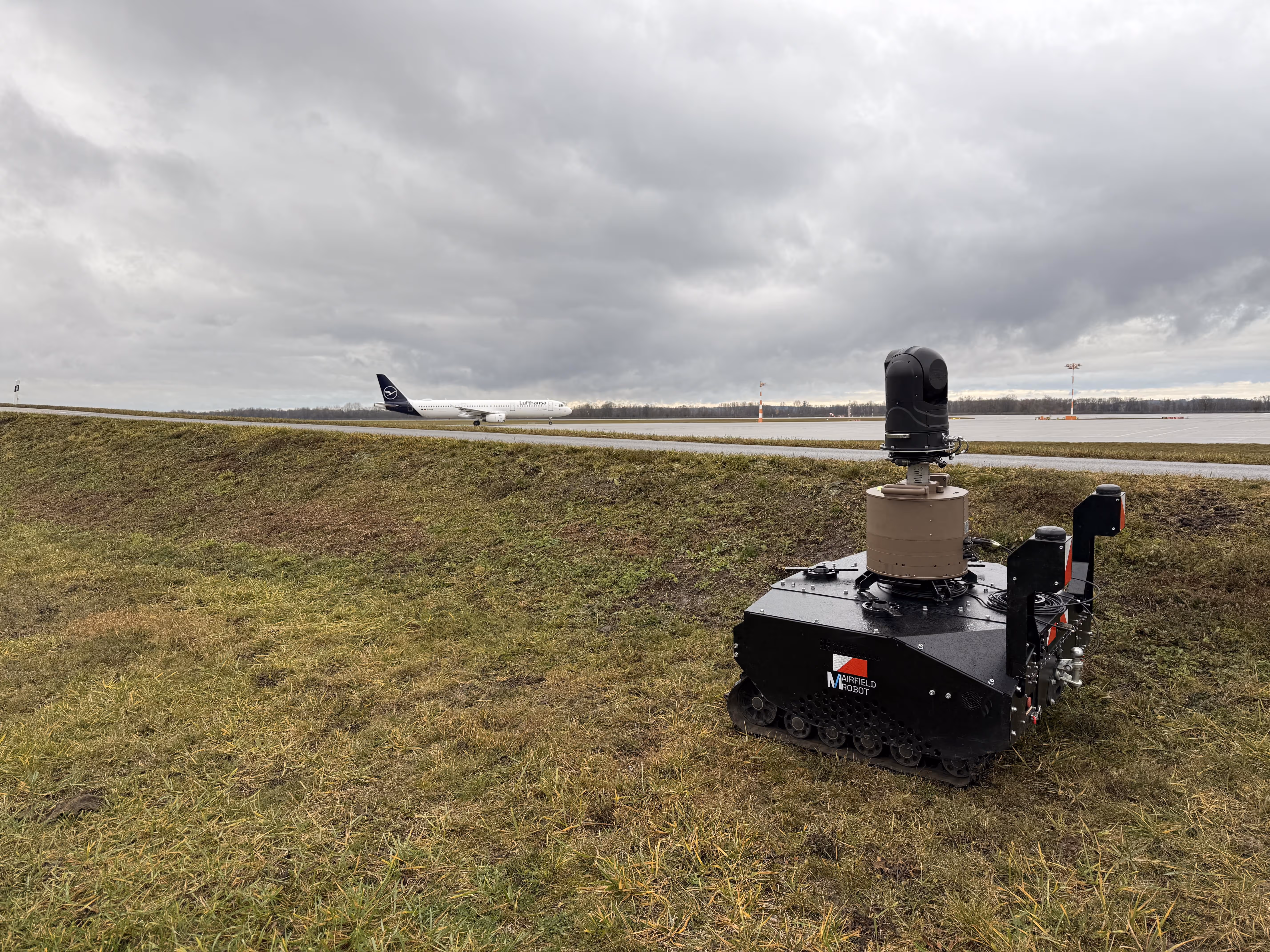Black tracked autonomous robot with sensors on grass near an airport runway with a Lufthansa airplane taxiing in the background under a cloudy sky.