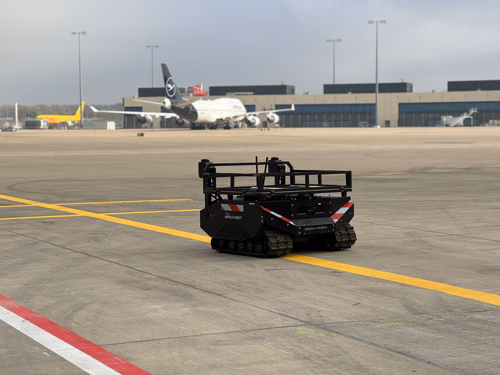 Small black tracked robot labeled 'Airfield Robot' on an airport tarmac with a Lufthansa airplane and airport terminal in the background.