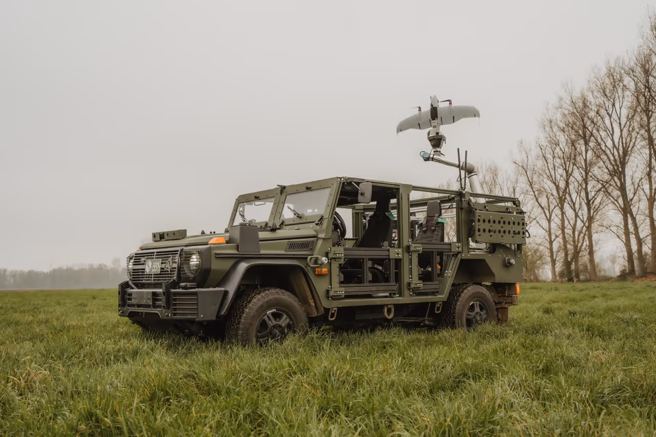 Military green unmanned ground vehicle parked on grass with a drone mounted on an arm above it.