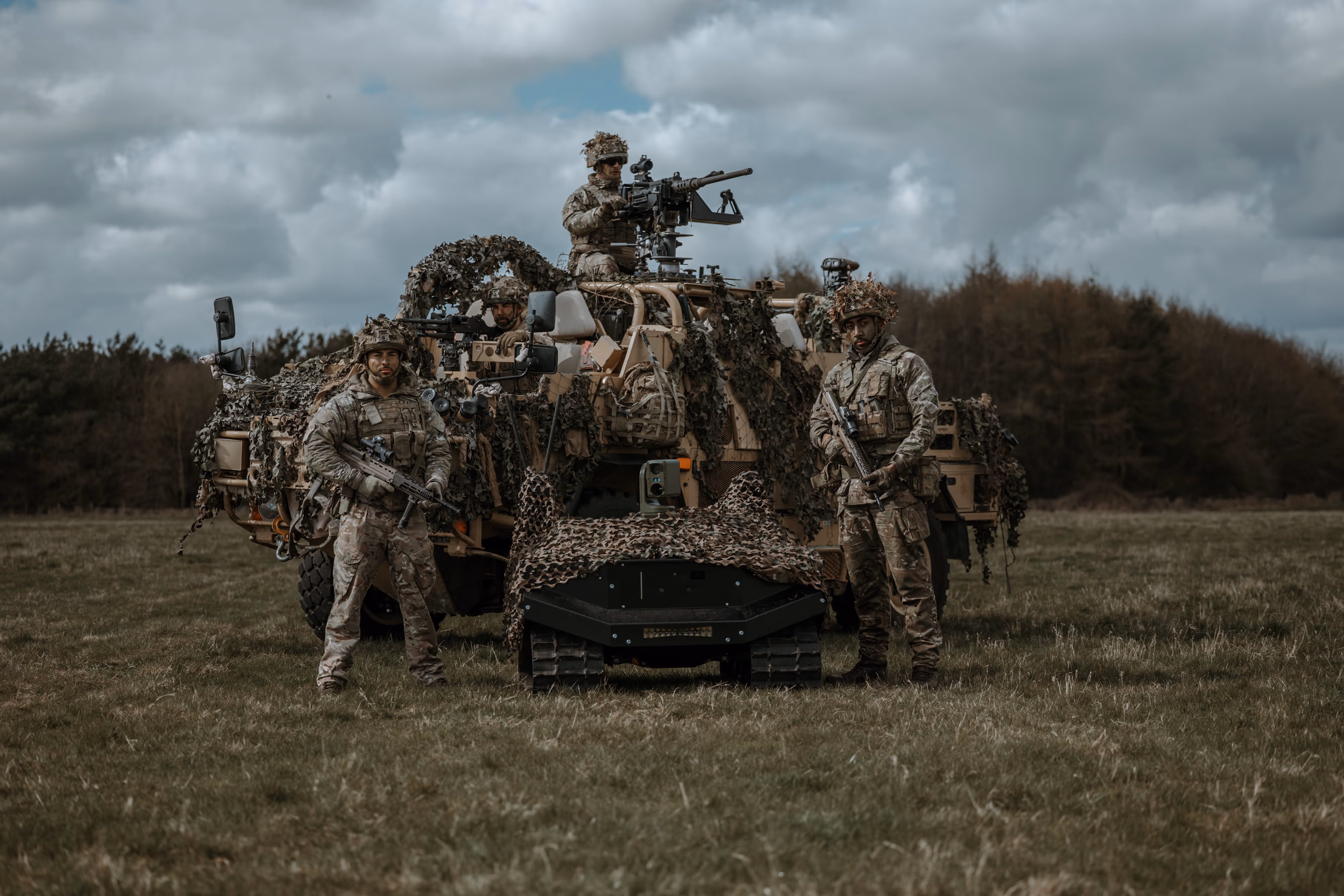 Four soldiers in camouflage gear with rifles standing near a camouflaged military vehicle and a small tracked robot in an open field under a cloudy sky.