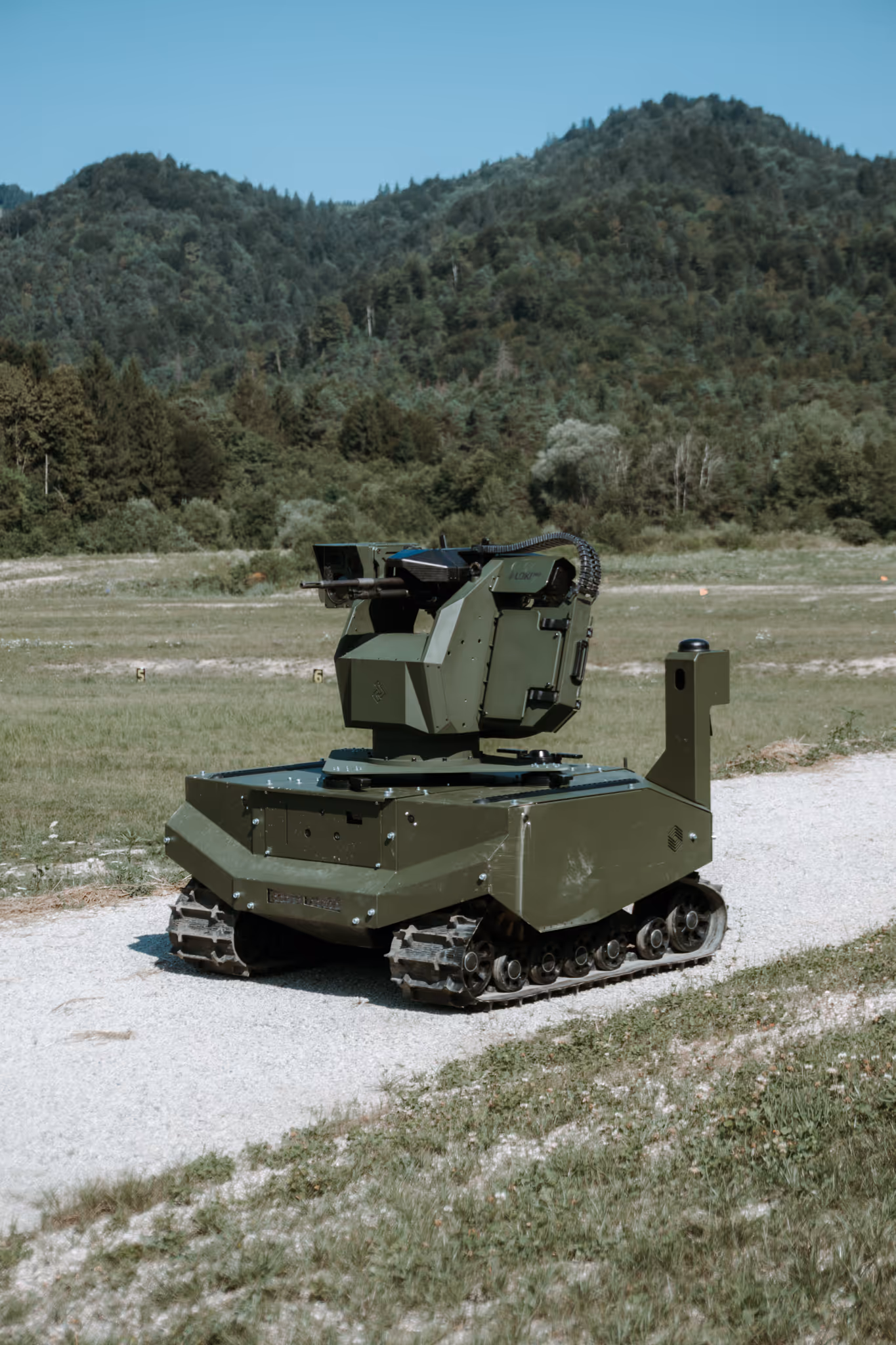 Military green combat unmanned ground vehicle with mounted weapon on a gravel path in a grassy field with forested hills in the background.