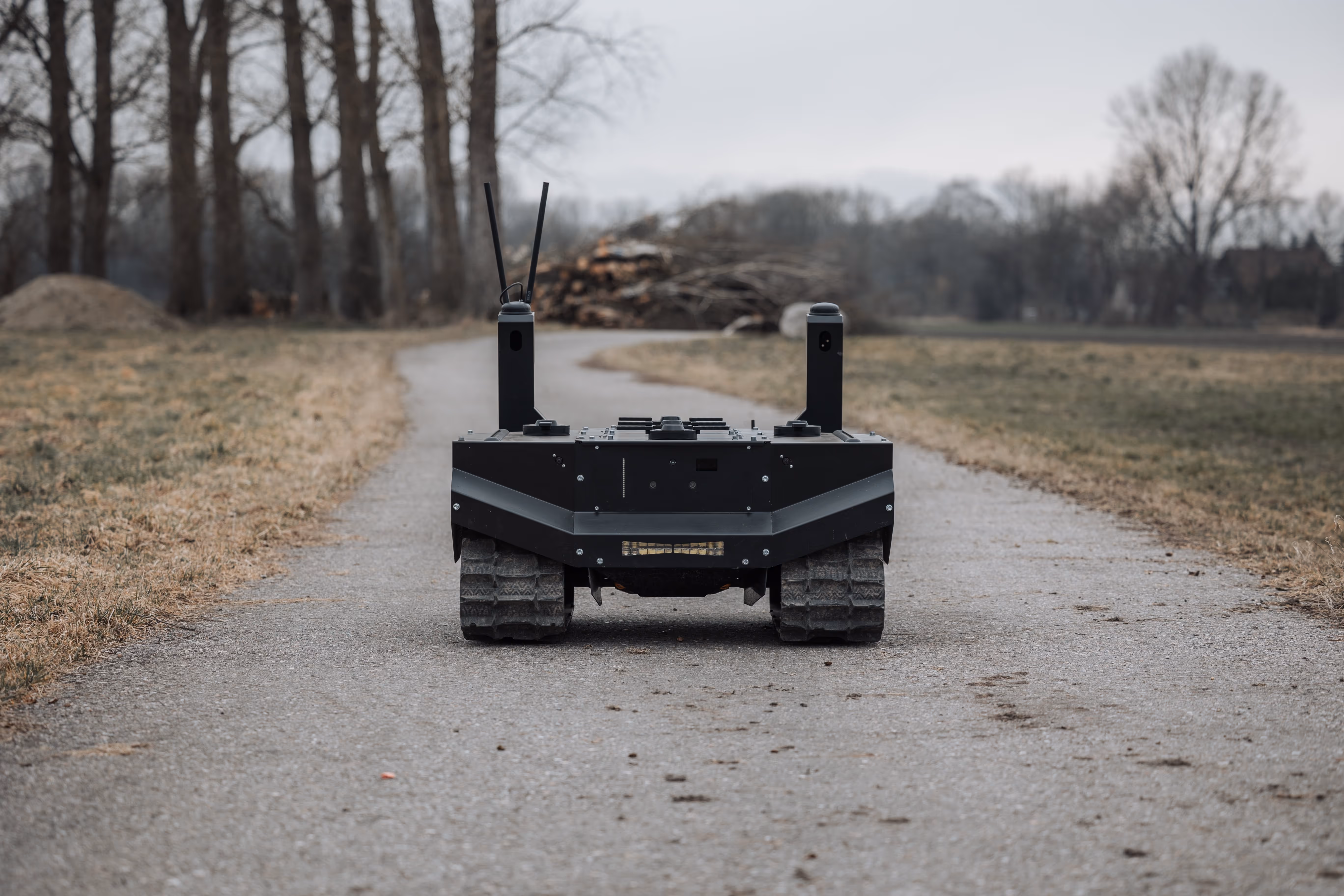 Black tracked robotic vehicle on a gravel path in a brown and green field with leafless trees in the background.