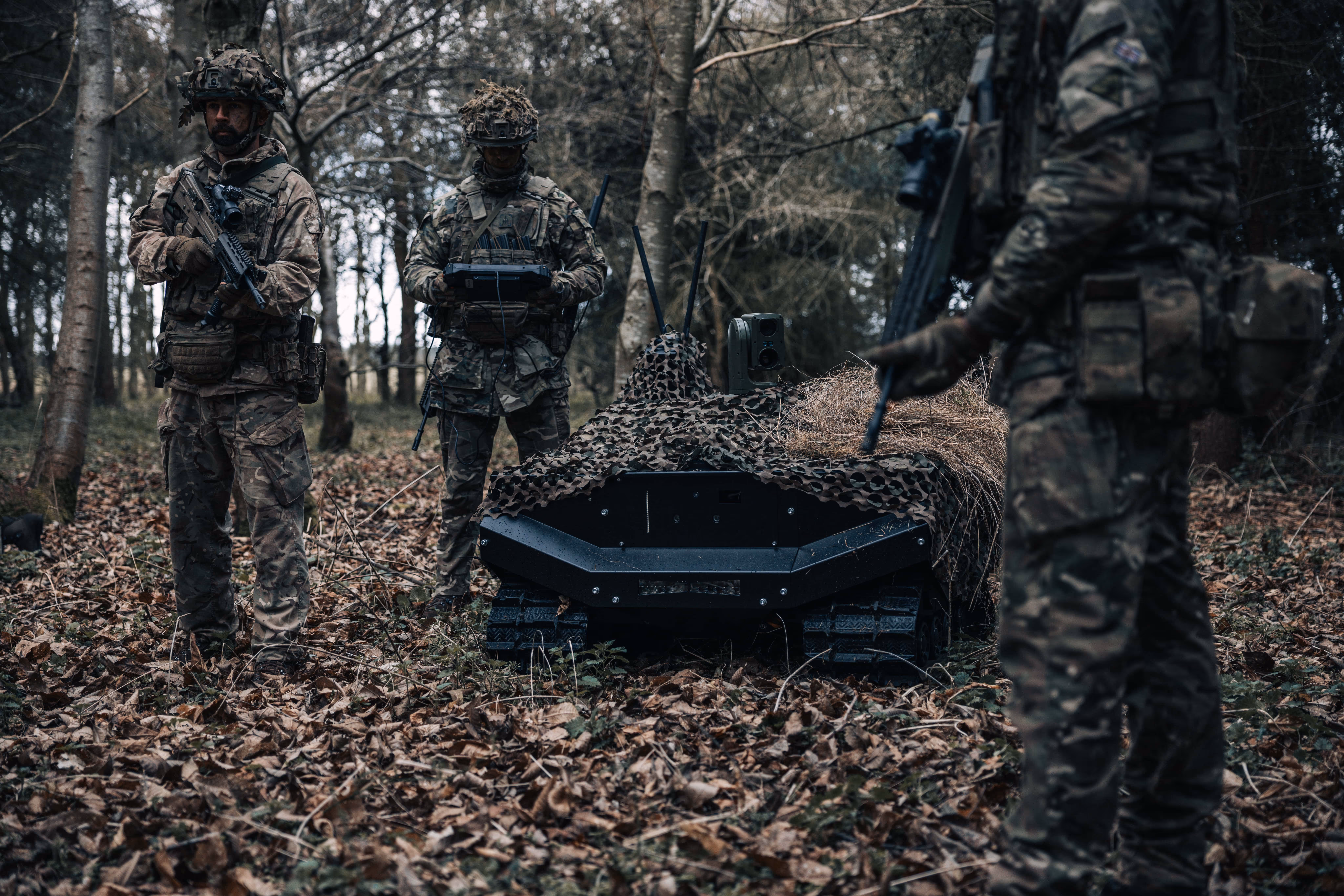 Three soldiers in camouflage gear stand in a forest beside a camouflaged robotic tracked vehicle.