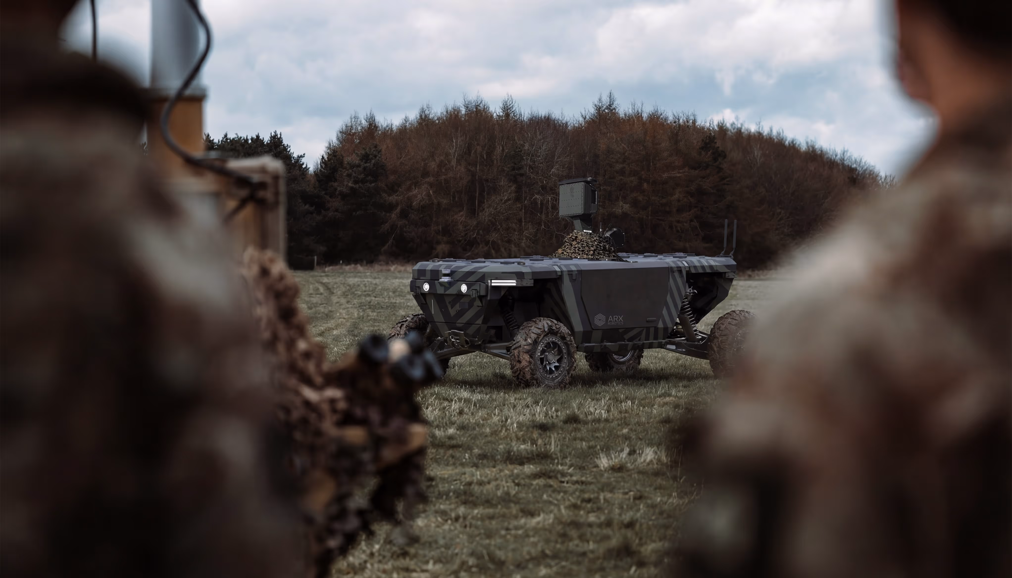 Camouflaged military autonomous vehicle on grassy field with two soldiers blurred in foreground.