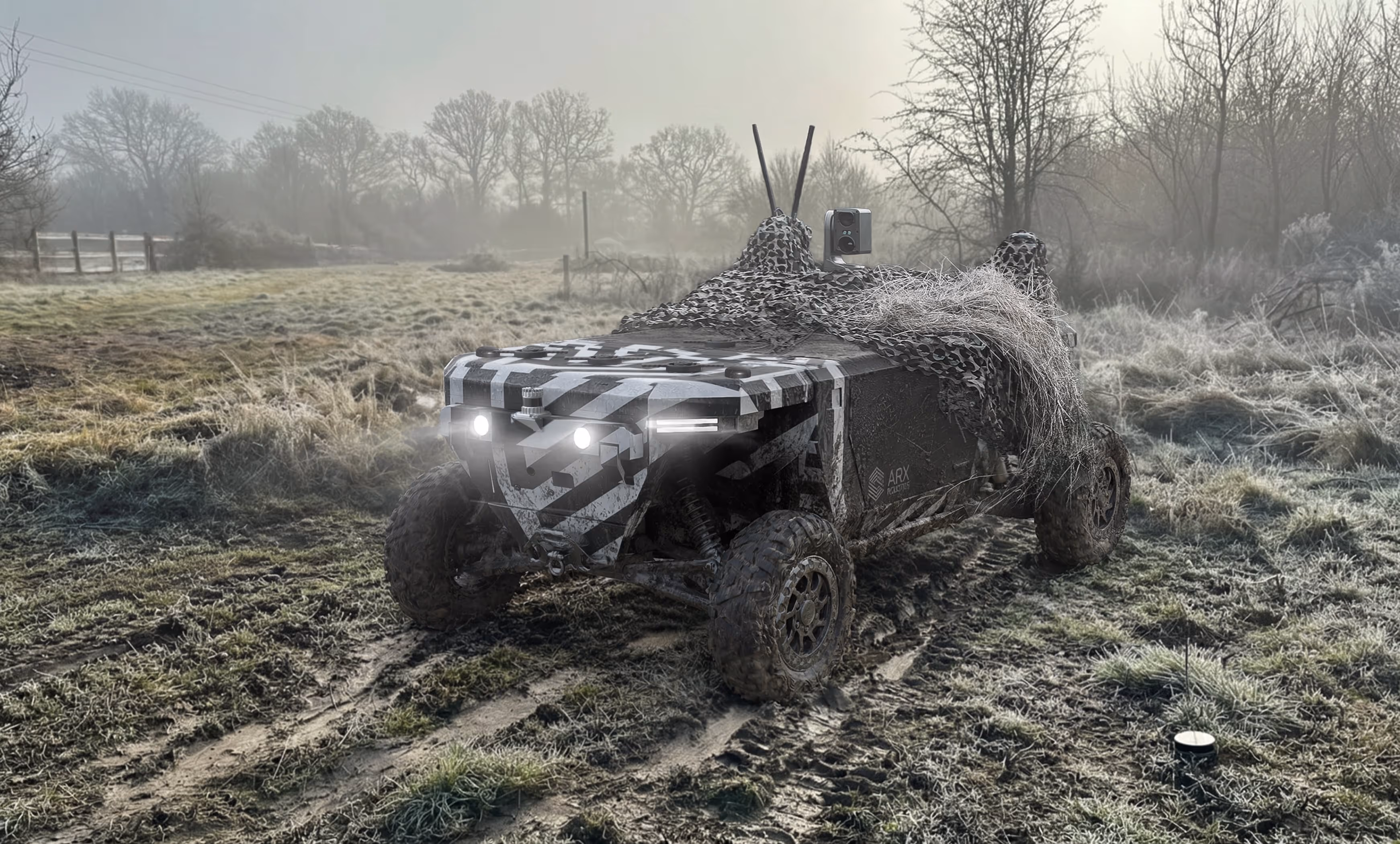 All-terrain camouflage vehicle with headlights on, driving on a muddy path in a frosty rural landscape.