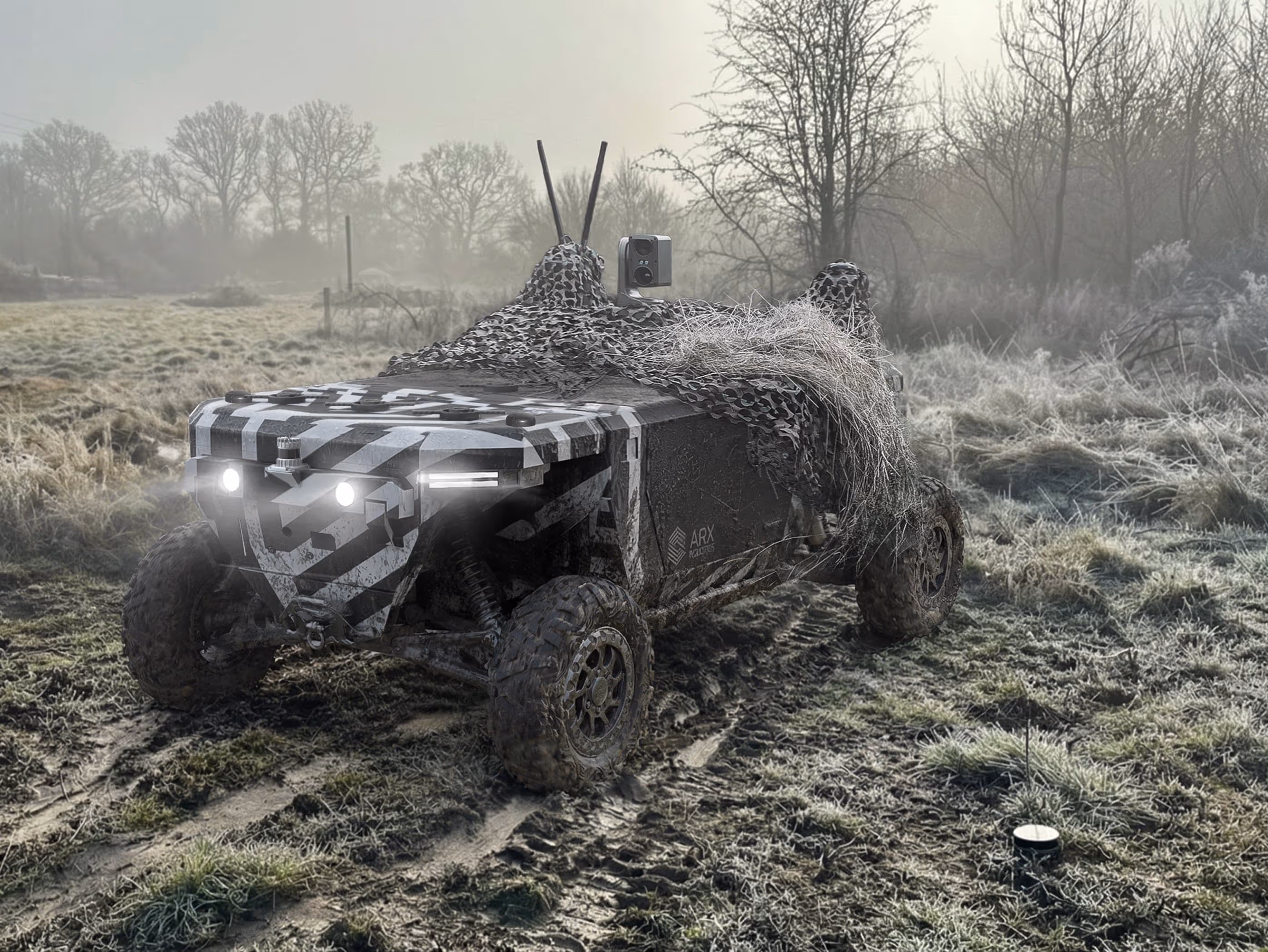 Camouflaged all-terrain vehicle with headlights on parked on muddy grass in a foggy rural area.