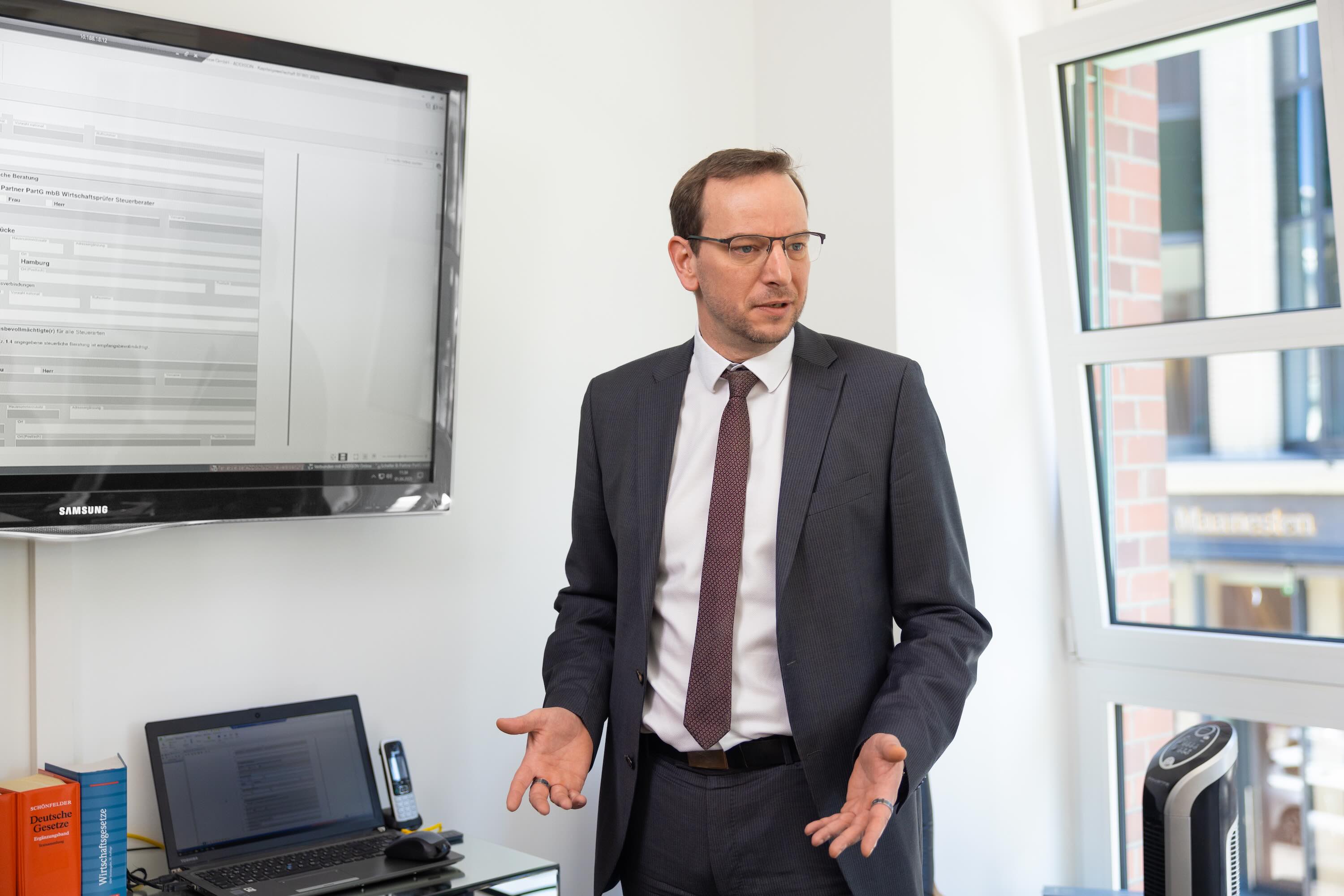 Mann im Anzug steht vor Präsentationsbildschirm, spricht.