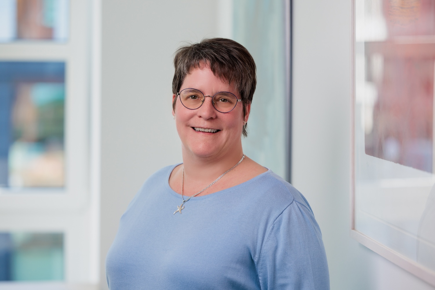Porträt einer Frau mit Brille im hellblauen Oberteil, lächelnd.