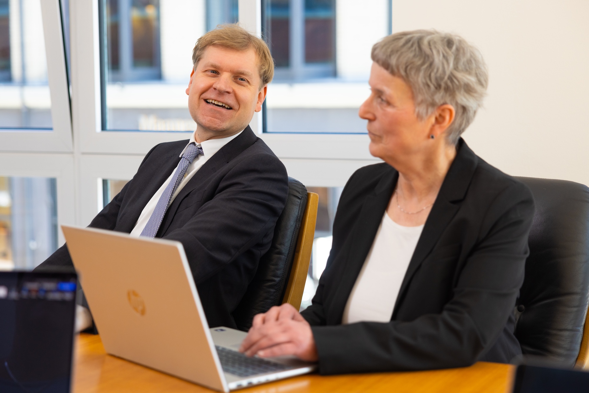 Zwei Personen im Anzug sitzen am Tisch mit Laptop, sprechen miteinander.