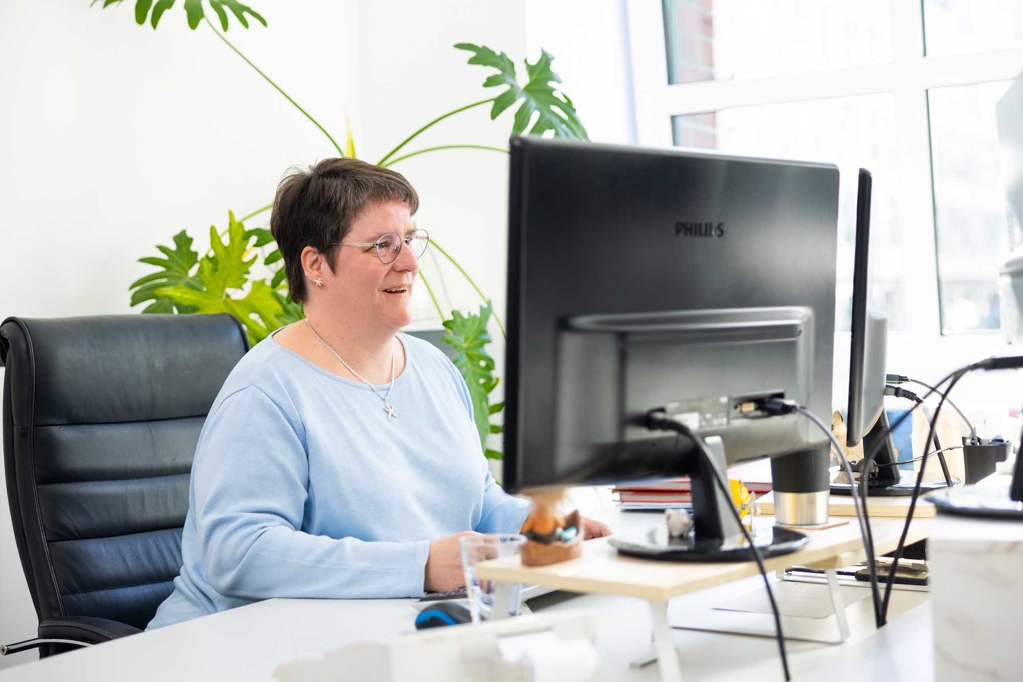 Frau sitzt am Schreibtisch vor PC, lächelnd.