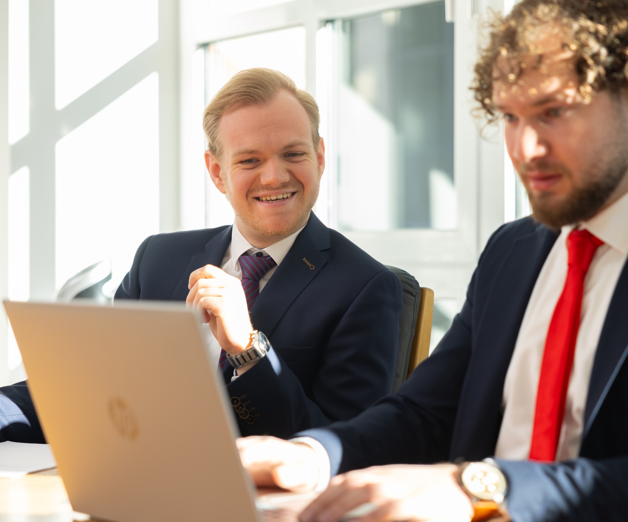 Zwei Männer im Anzug lächeln, arbeiten gemeinsam am Laptop.