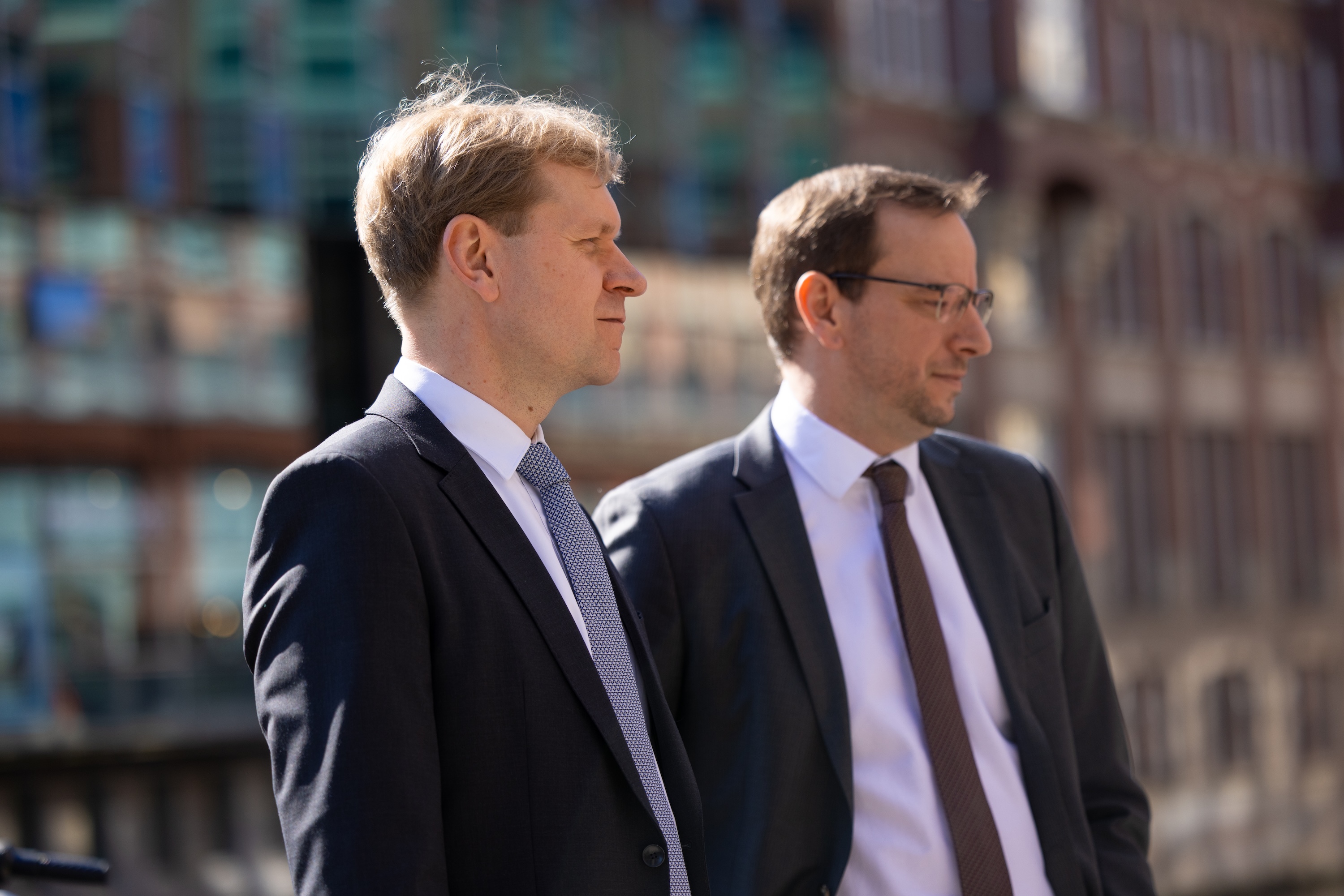 Zwei Männer im Anzug gehen nebeneinander draußen im Gespräch.