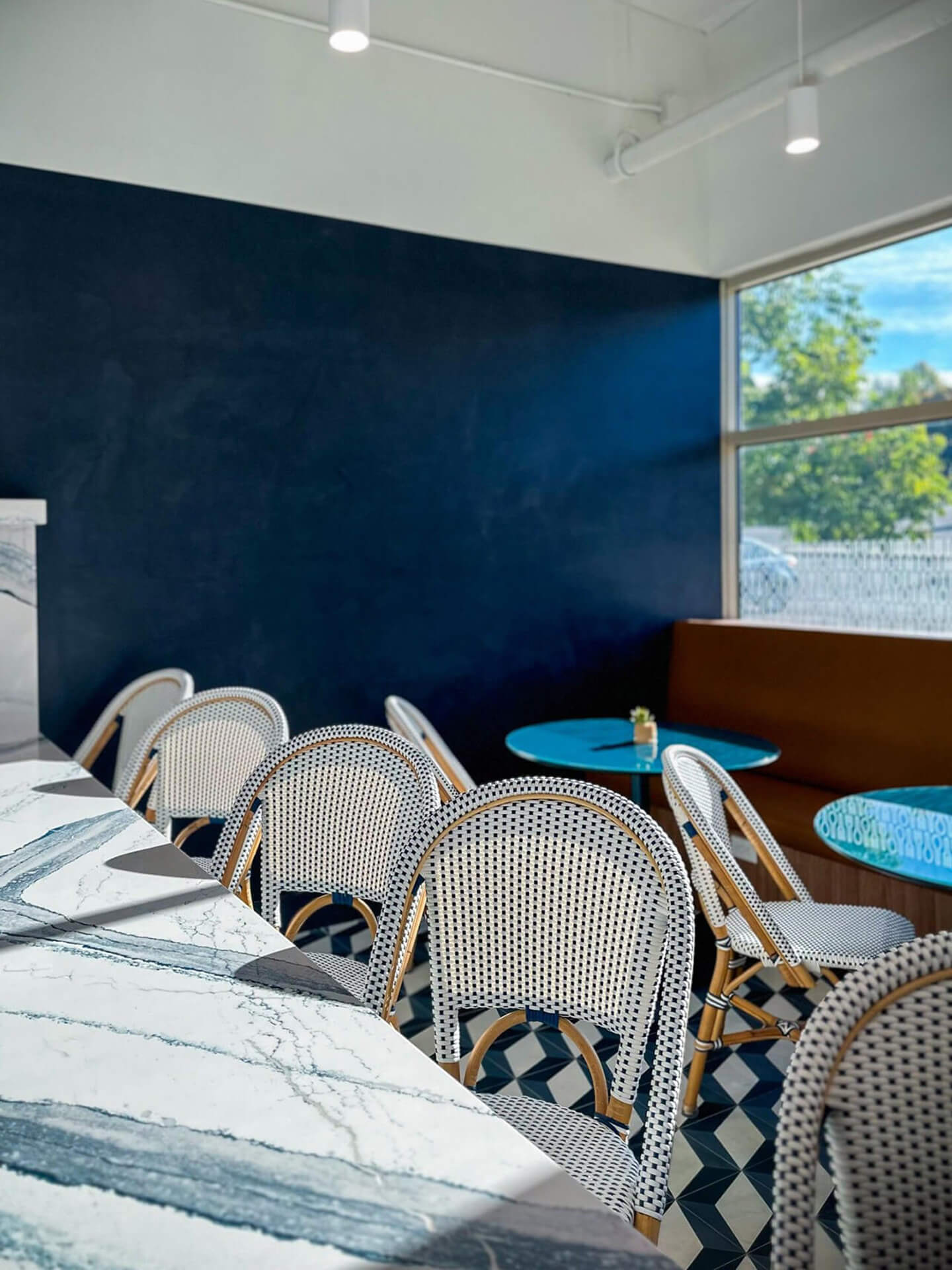 A modern break room interior with white wicker bistro chairs, marble tables, blue accent walls, geometric floor tiles, and a window letting in natural light with trees visible outside.