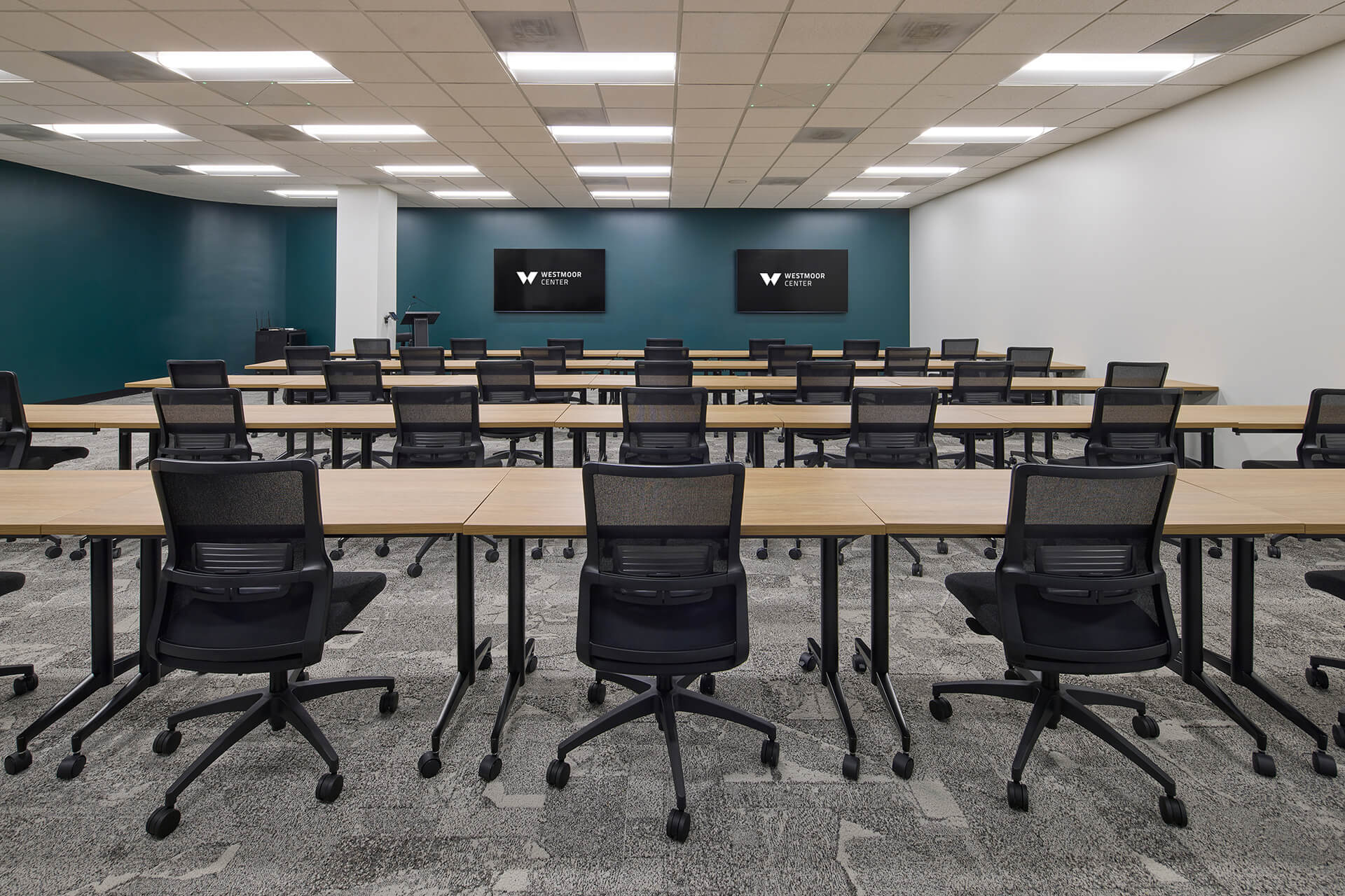 A modern classroom with rows of empty desks and black chairs facing two large screens on the front wall; teal and white walls, gray carpet, and ceiling lights create a clean, professional setting.