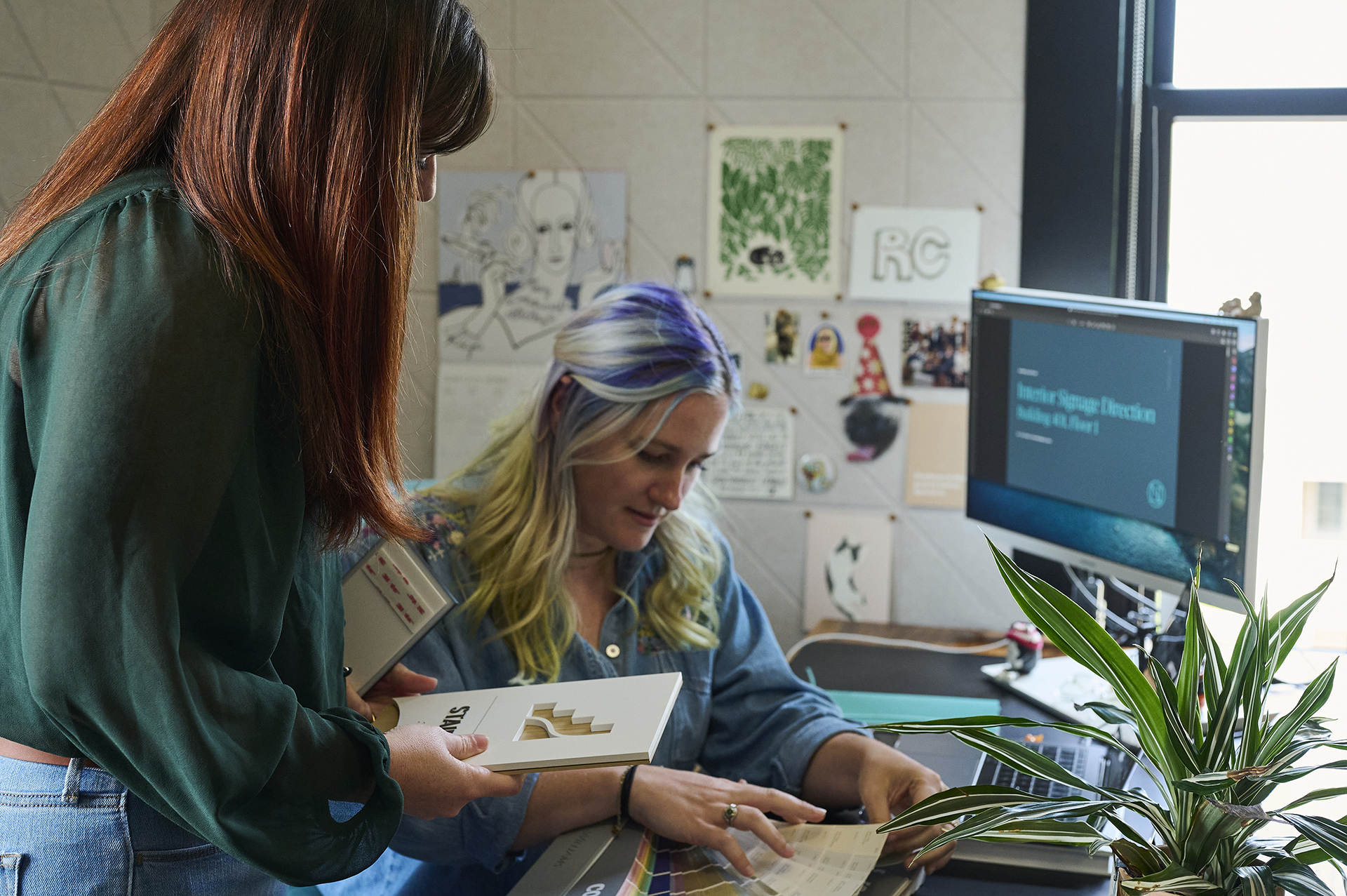 Two women work together at a desk; one is seated looking at documents, while the other stands holding a notebook. A computer monitor, plant, and artwork are visible in the background.