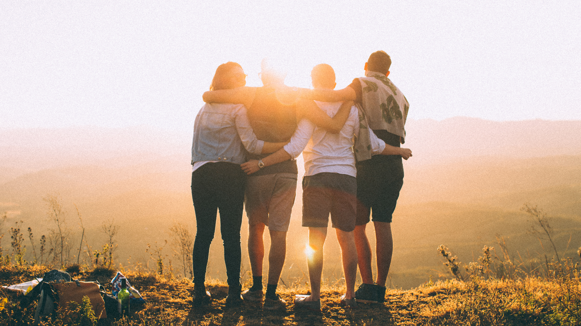 Four friends standing on mountain top looking into the sun, embracing each other