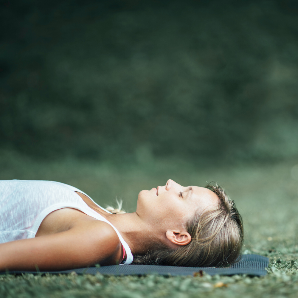 Woman seen in profile laying down on yoga mat in the grass with closed eyes looking serene