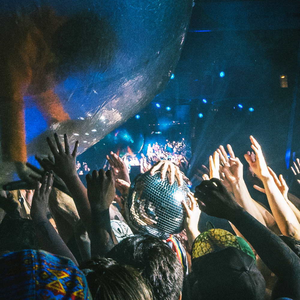 Disco indoors with hands reaching upwards and a disco ball in the center being held by a pair of hands