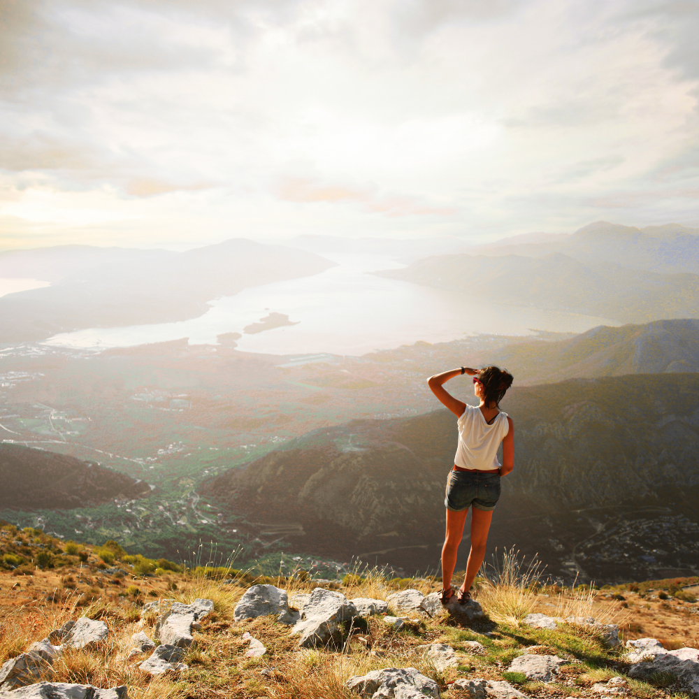 Woman from behind standing at a cliff looking out over scenery with mountains, valleys and water in the sunshine