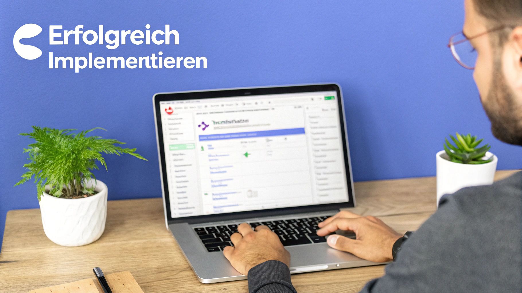 Person tippt am Laptop auf einem Holztisch vor einer blauen Wand mit dem Schriftzug 'Erfolgreich Implementieren' und Pflanzen.