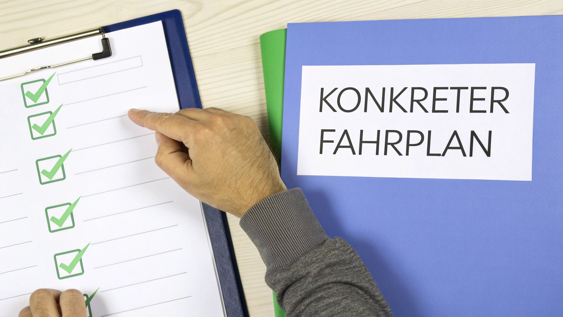Eine Hand zeigt auf eine Checkliste mit grünen Häkchen, daneben liegt ein blauer Ordner mit der Aufschrift 'Konkreter Fahrplan'.
